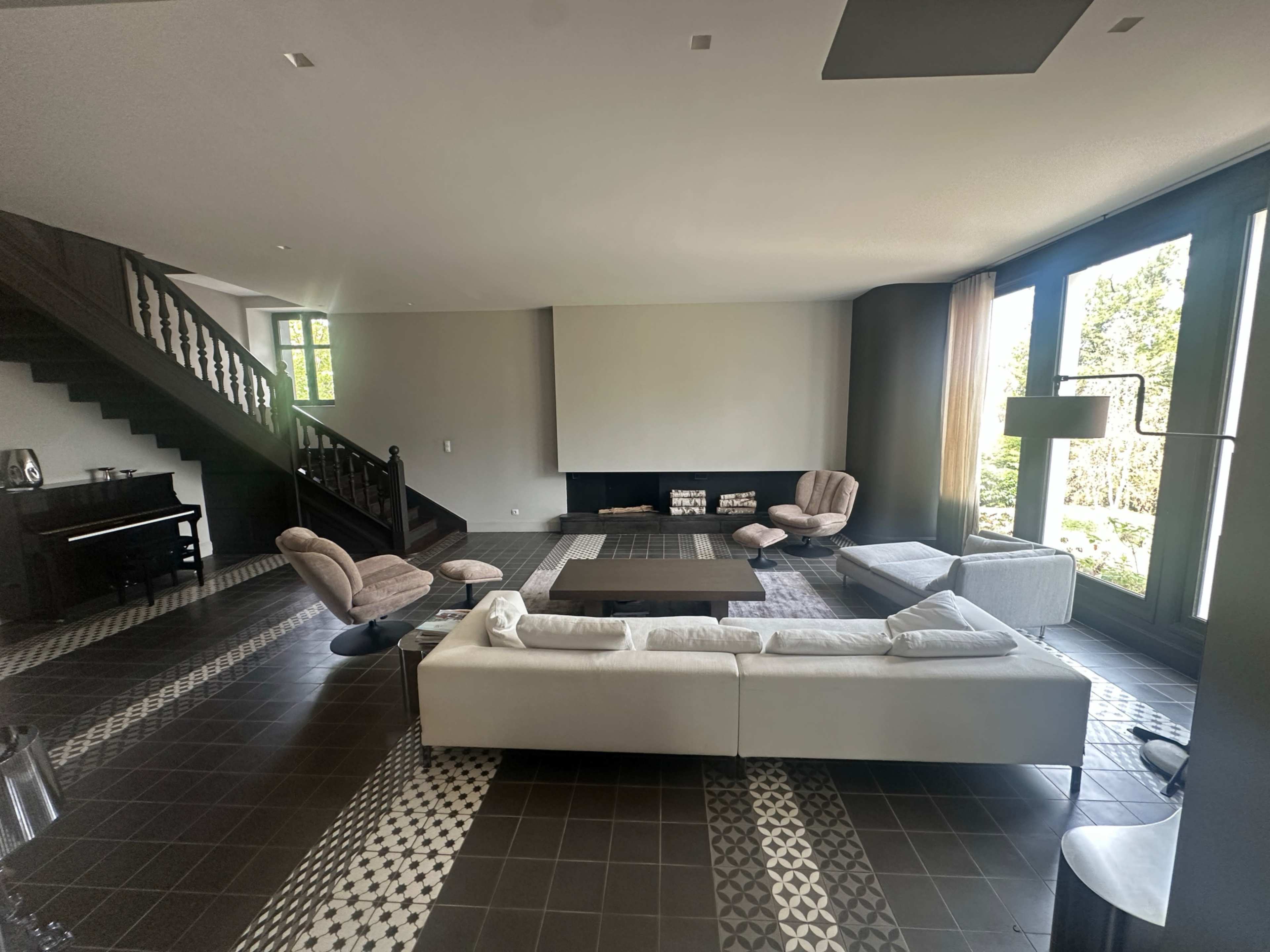 The image shows a spacious, modern living room with large windows, a light-colored sofa, a coffee table, and various seating arrangements, including armchairs and a small piano.