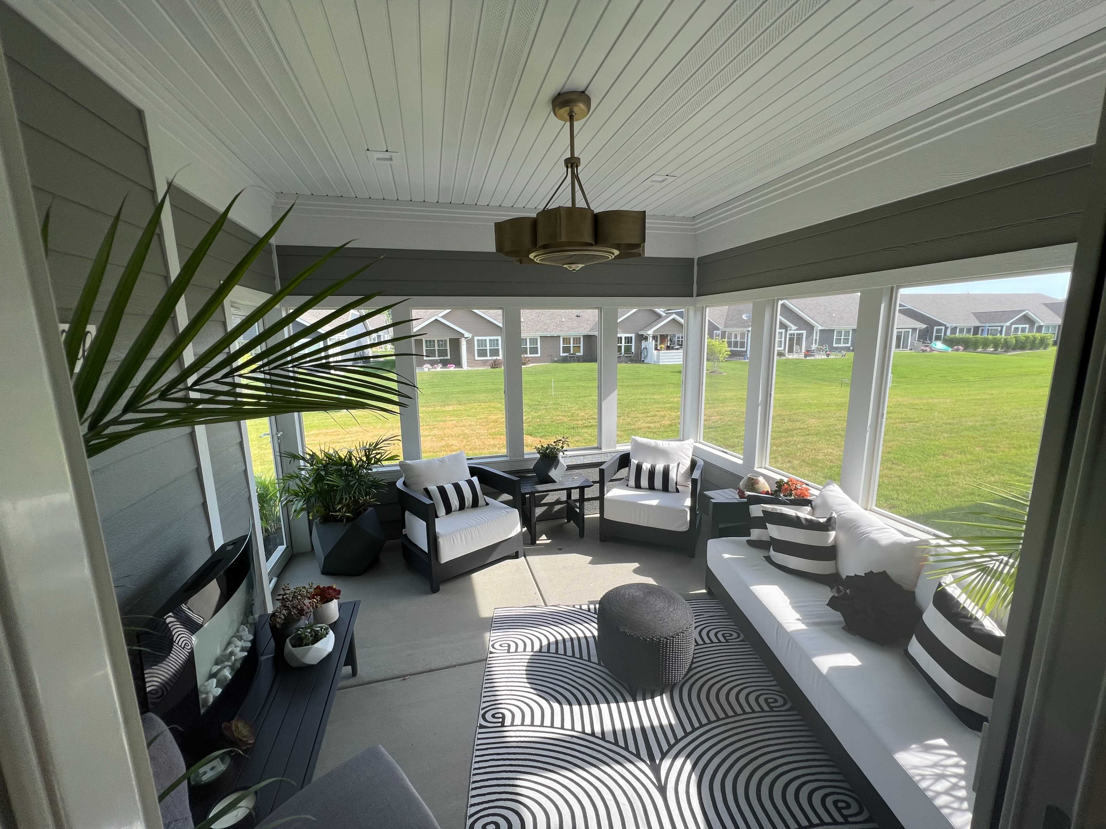 The image shows a sunroom with seating furniture, a round table, and indoor plants, featuring large windows overlooking a grassy area.