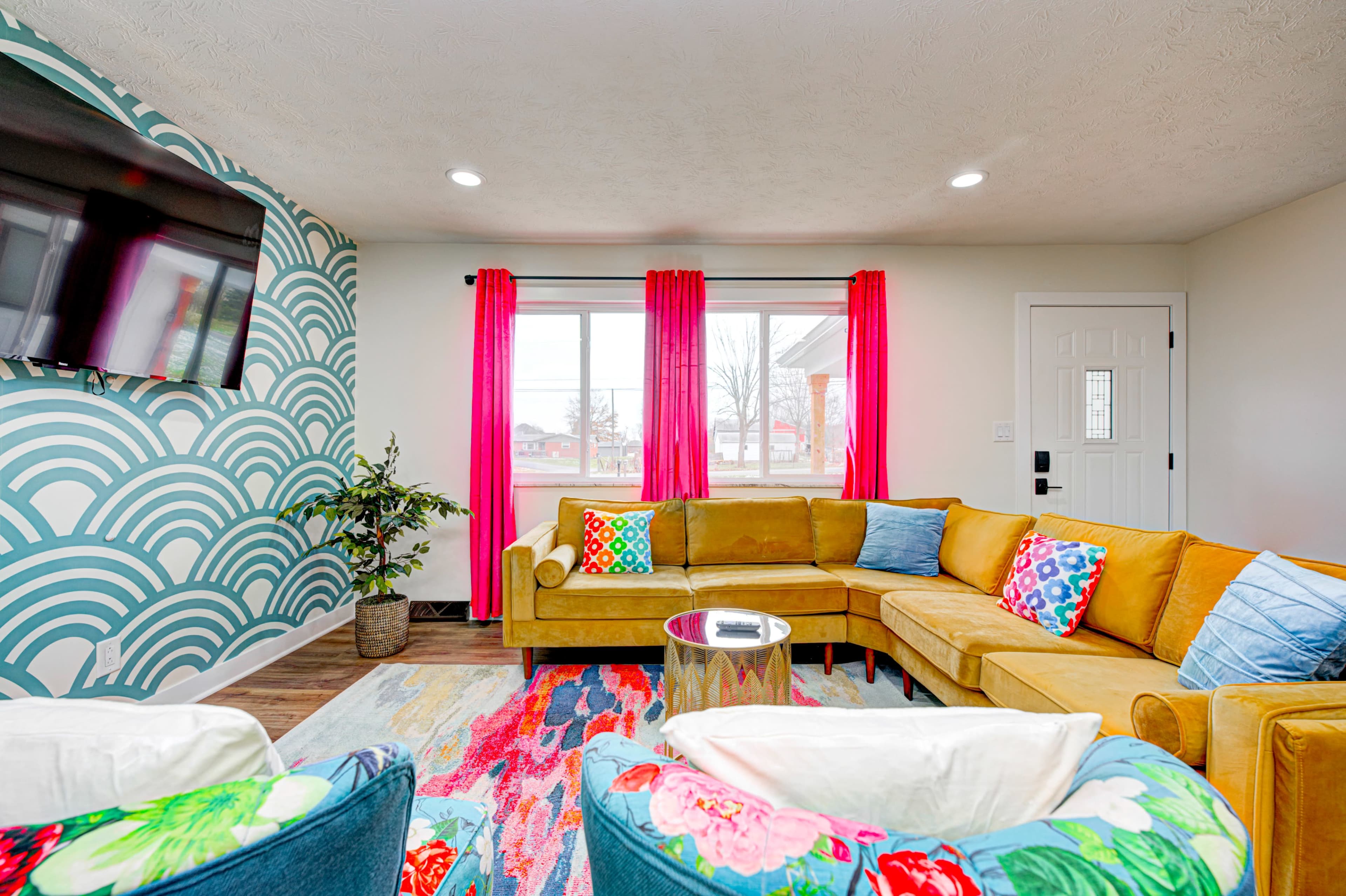 A brightly decorated living room features a large yellow sectional sofa, colorful throw pillows, a round side table, and a patterned rug, with large windows and a television on the wall.