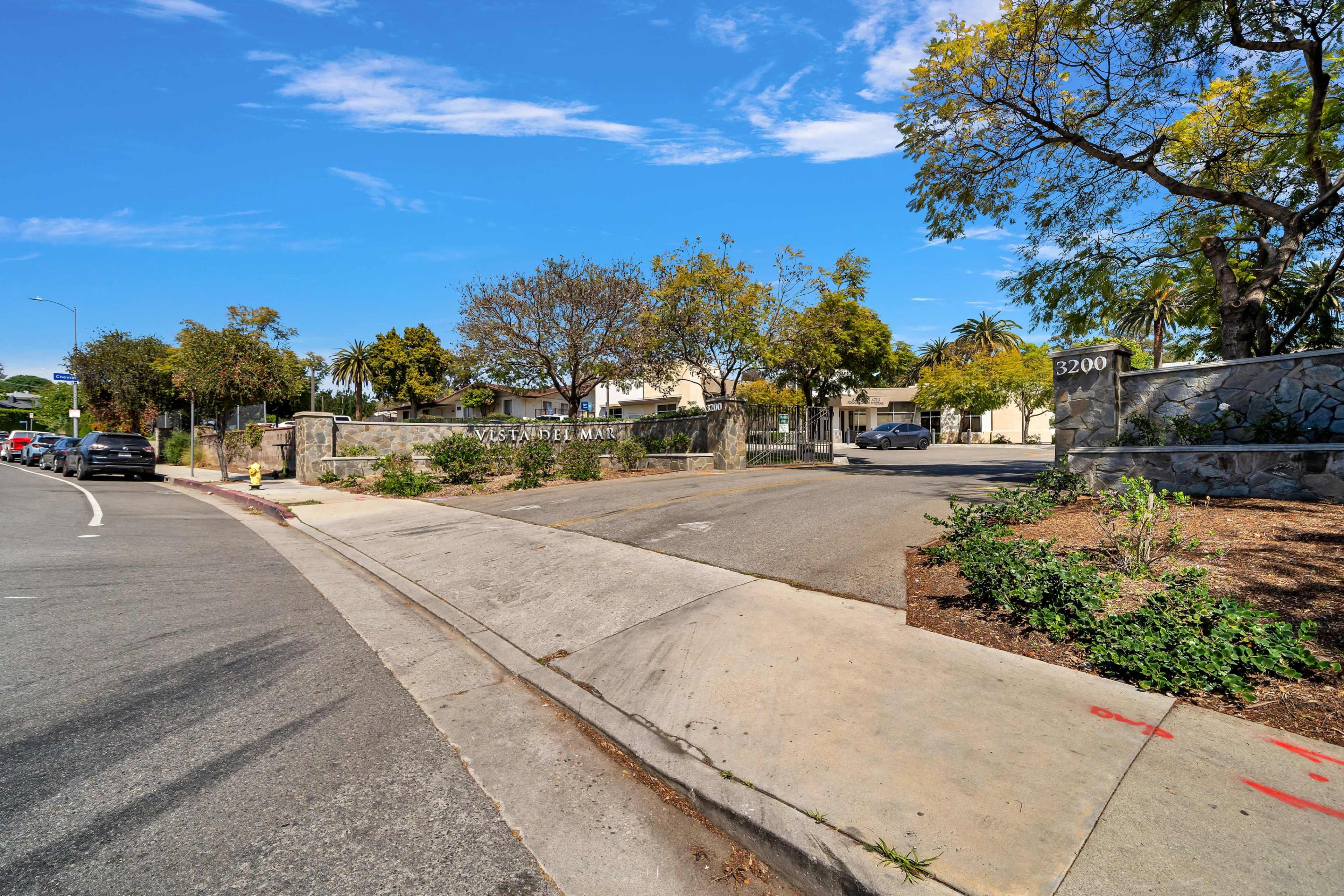 A road leading to a residential entrance marked by a stone wall and greenery.