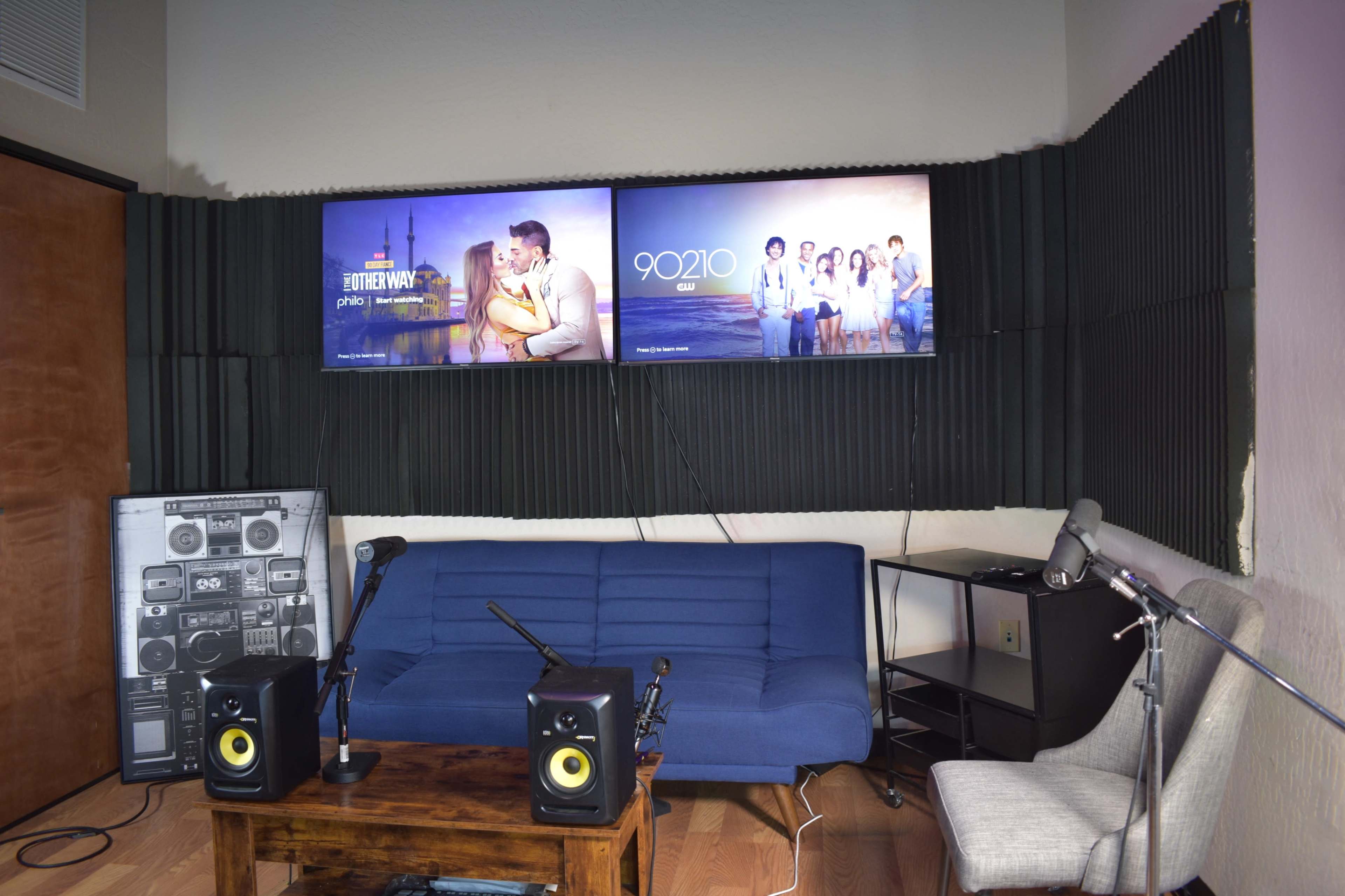 A room with two wall-mounted screens displaying TV show titles, a blue sofa, a wooden coffee table, and audio equipment.