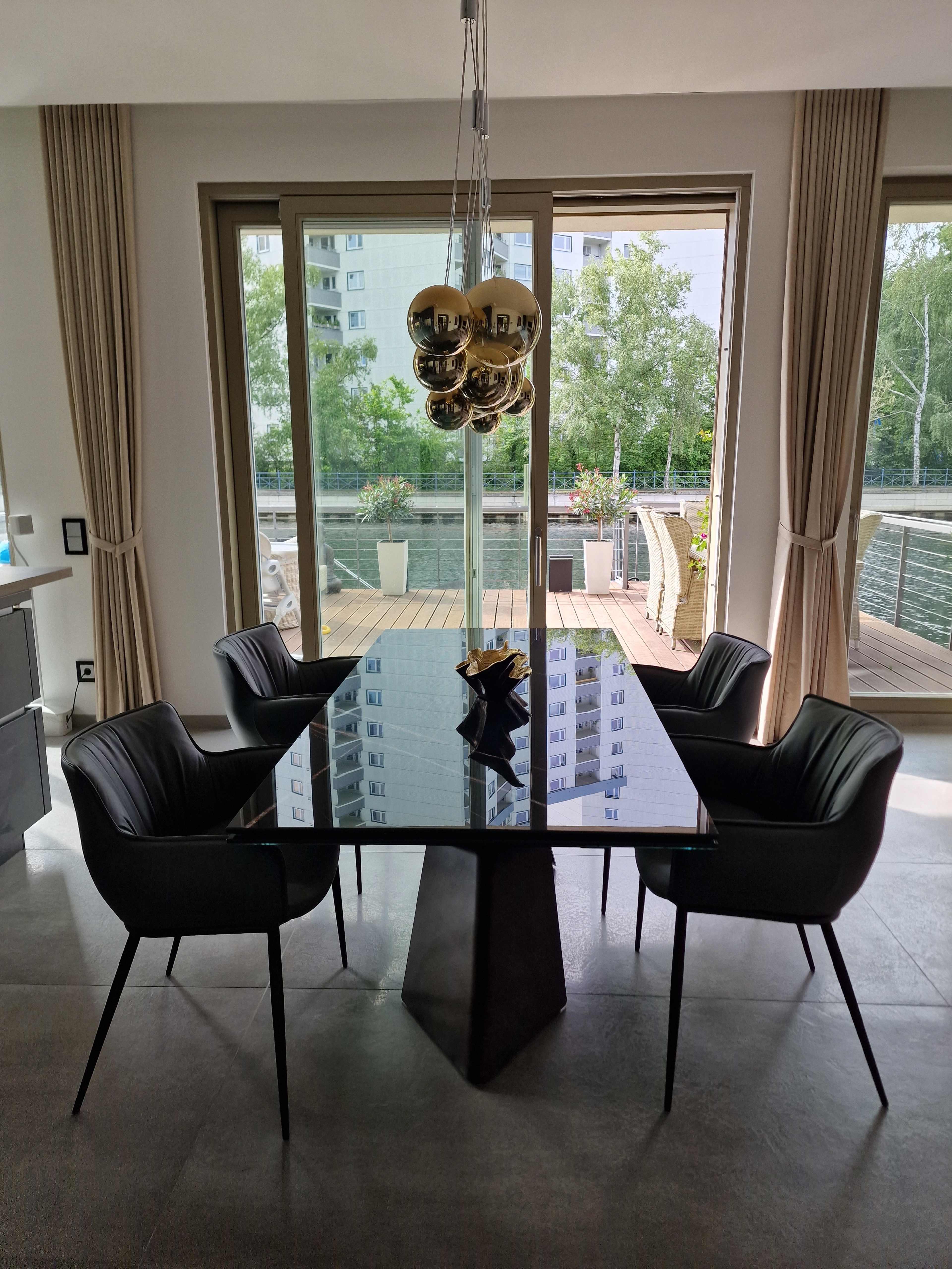 A modern dining area features a glass table with a geometric base surrounded by four black chairs, set near large windows overlooking a wooden deck and greenery.