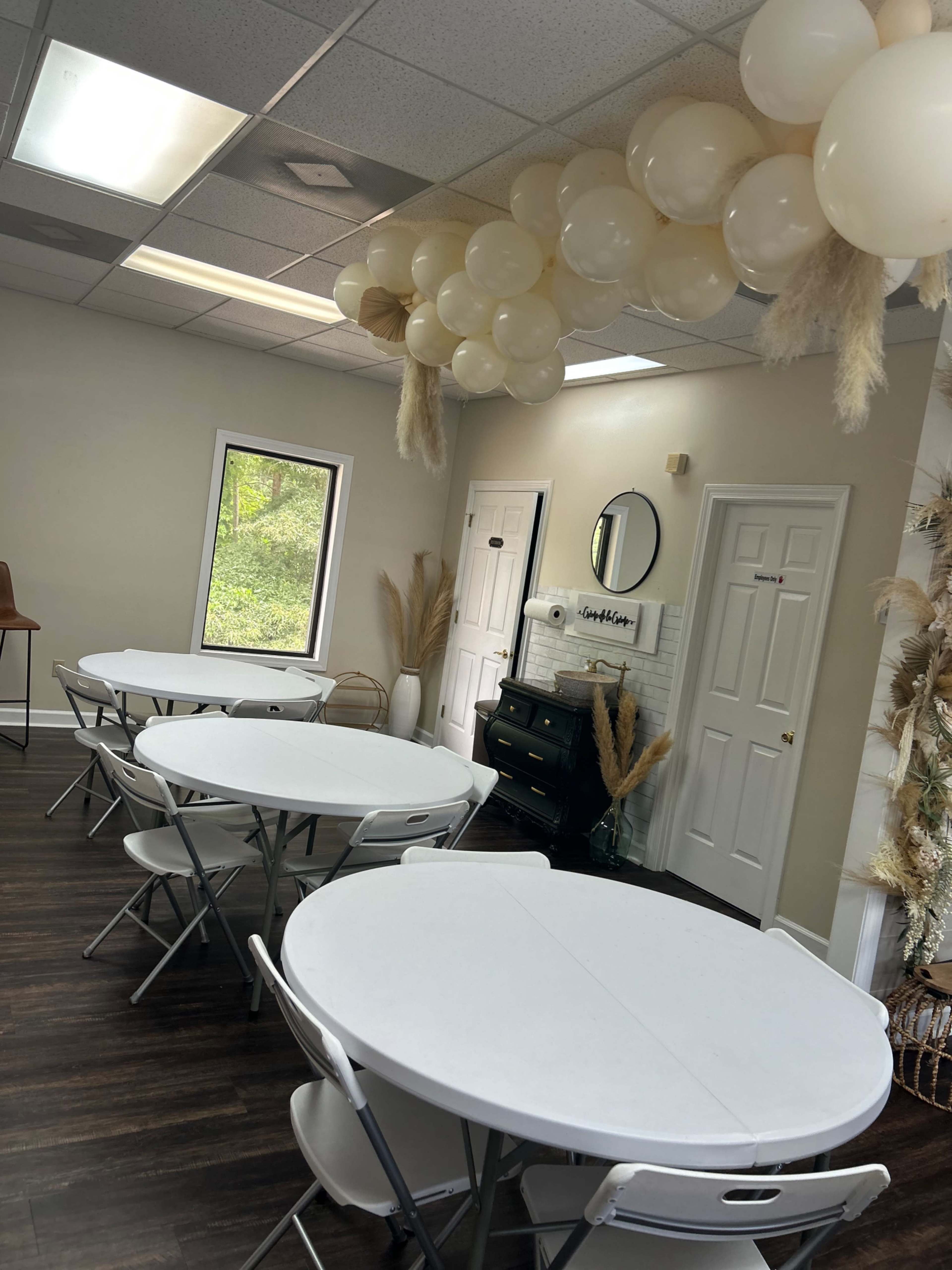 The image shows a lightly decorated event room with white circular tables, a faux fireplace, and a ceiling arrangement of pale balloons.
