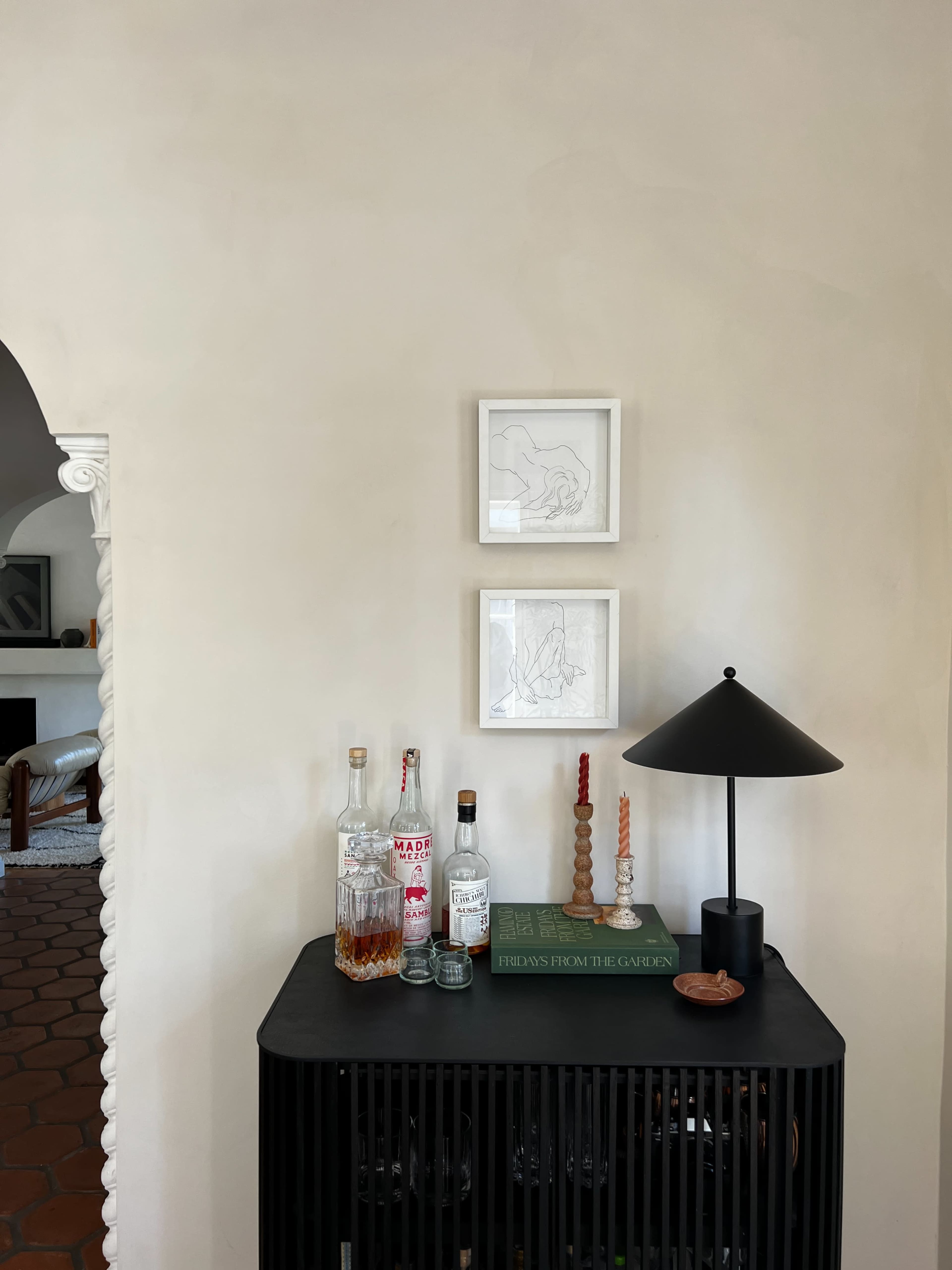 A minimalist black cabinet holds various bottles, a lamp, and books, with two framed artworks above it against a light-colored wall.