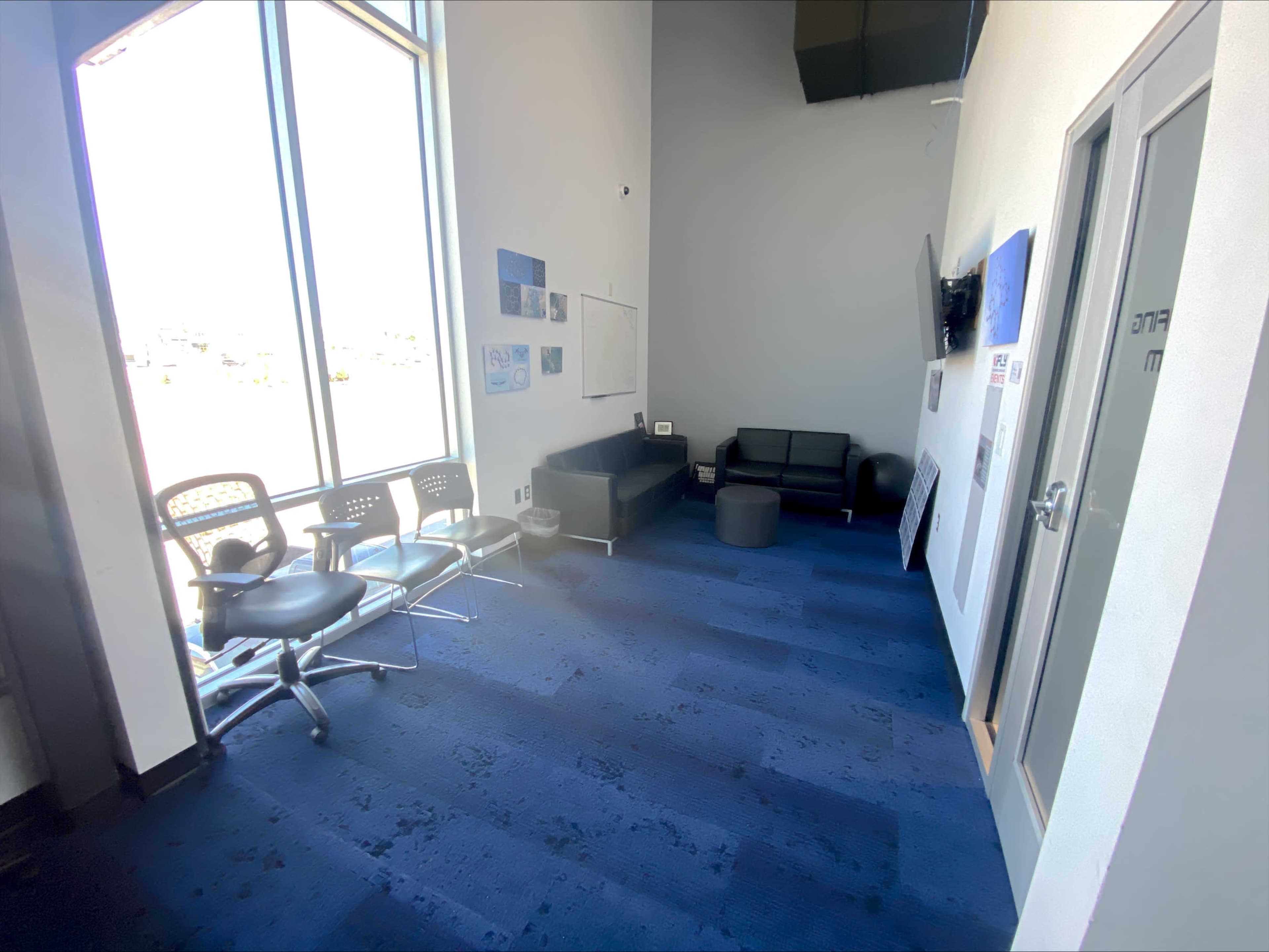A seating area with black chairs and couches arranged around a small table, located in a bright room with large windows and blue carpet.