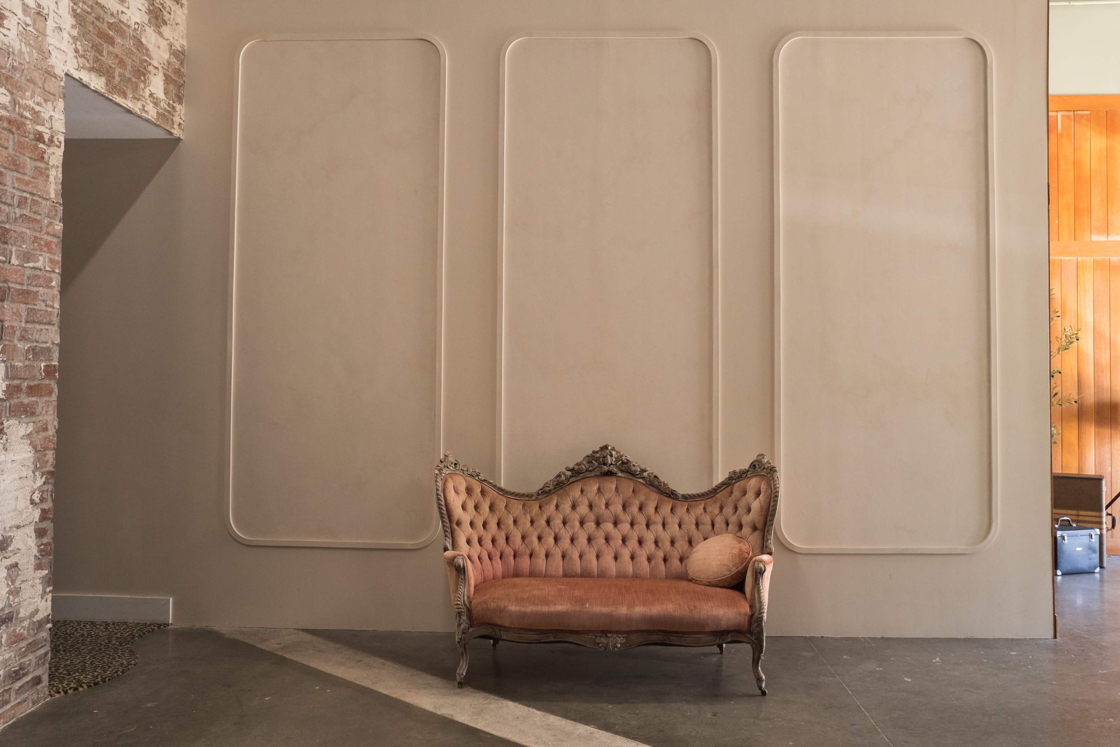 A vintage, tufted sofa with a decorative frame is positioned against a smooth, neutral-colored wall featuring three recessed panels.