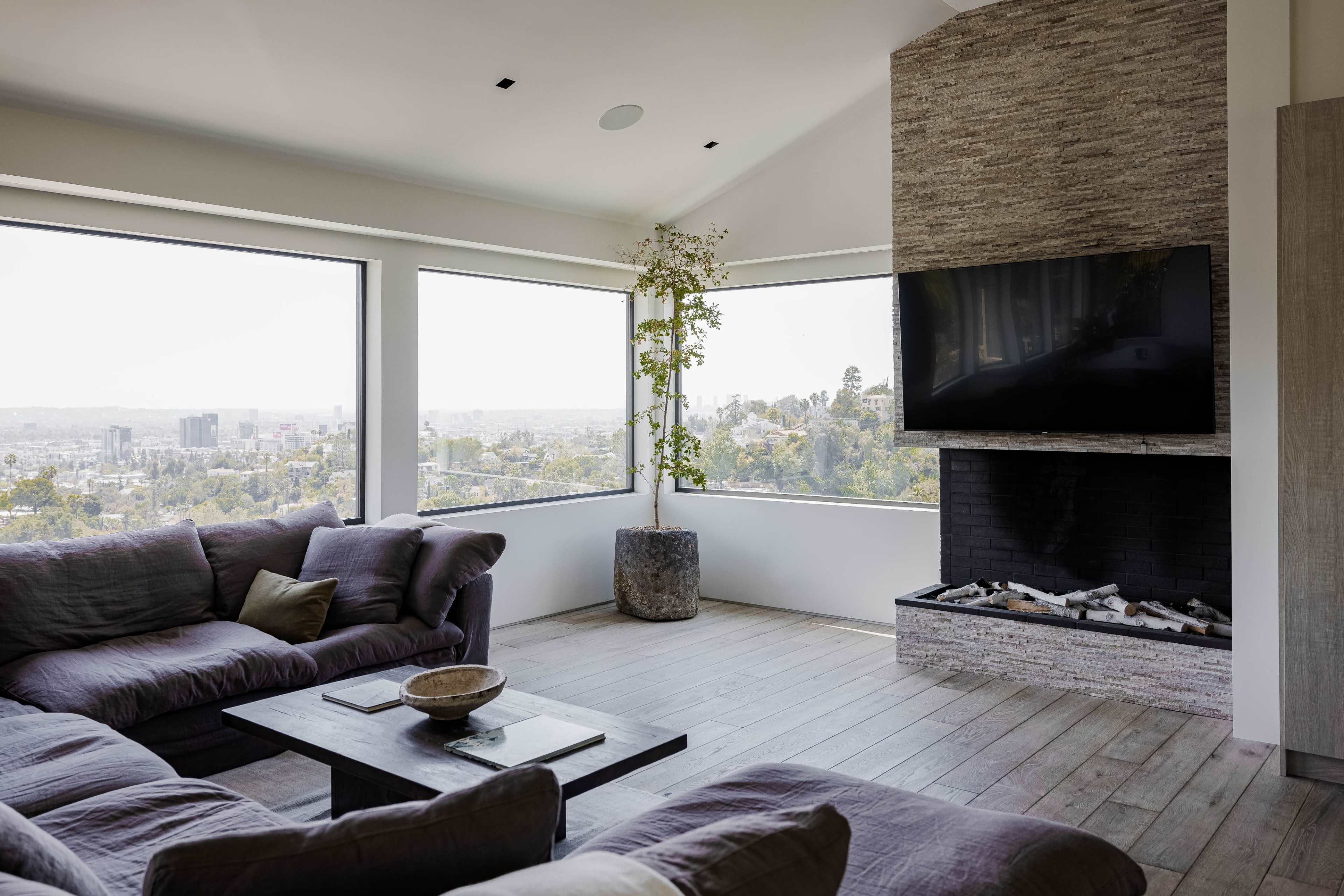 The image shows a modern living room with large windows, a flat-screen television, a stone fireplace, and a comfortable seating arrangement.