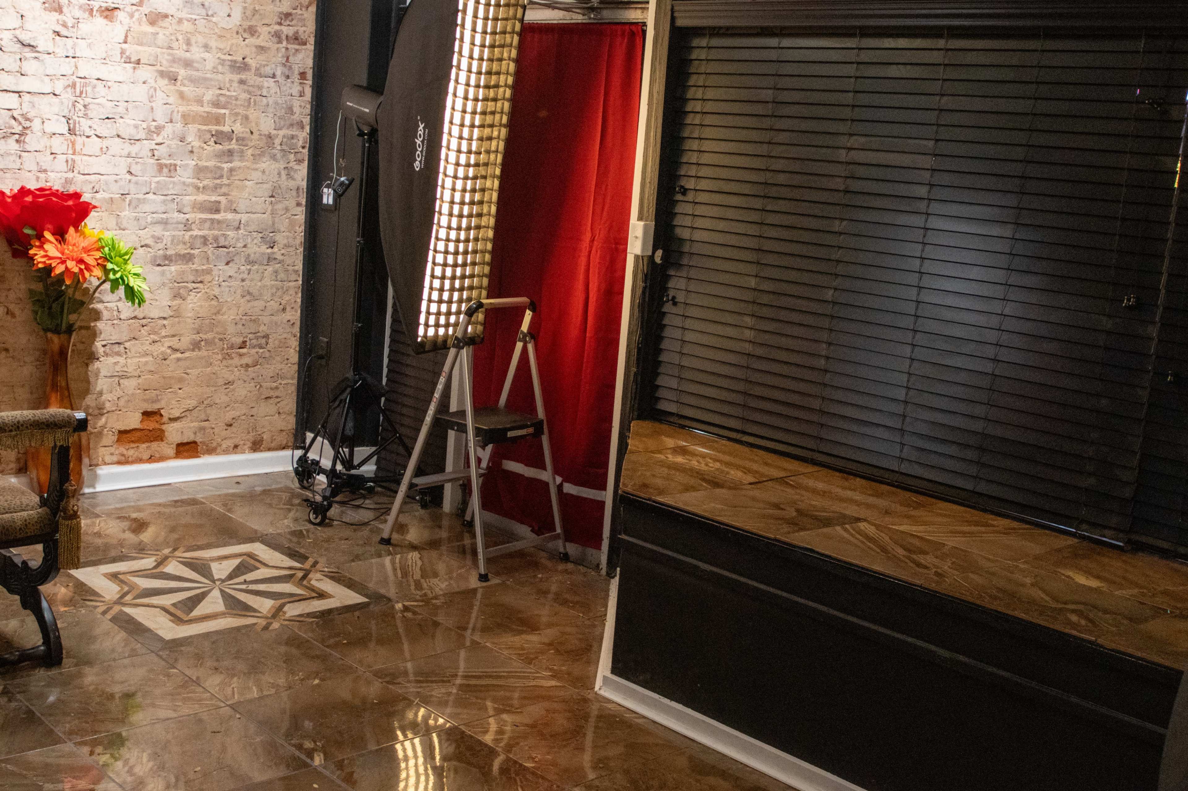 A room with a brick wall, a floral arrangement, a folding stepladder, and a backdrop with black blinds.