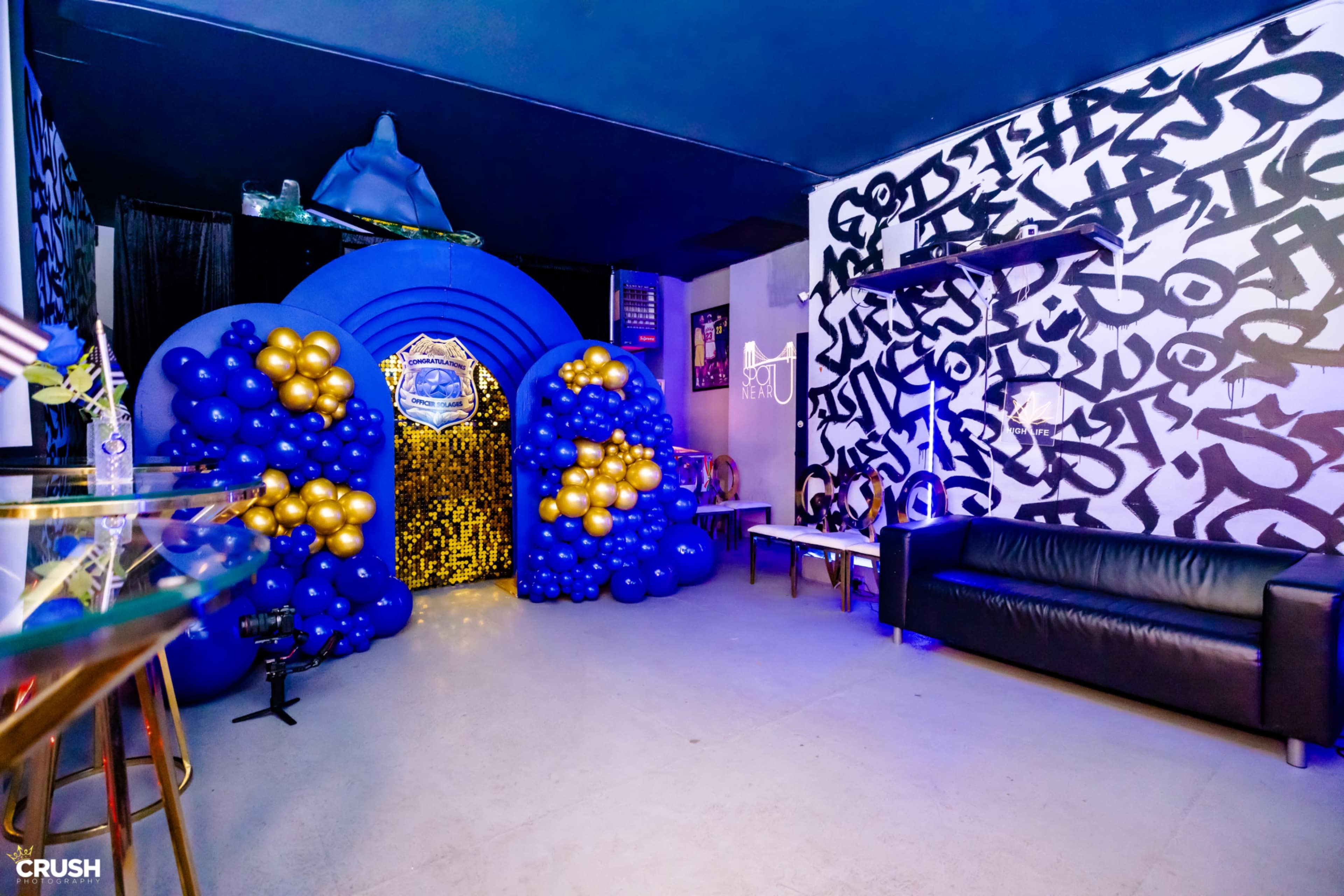 The image shows a vibrant party entrance decorated with blue and gold balloons, leading into a room with dark walls featuring graffiti art.