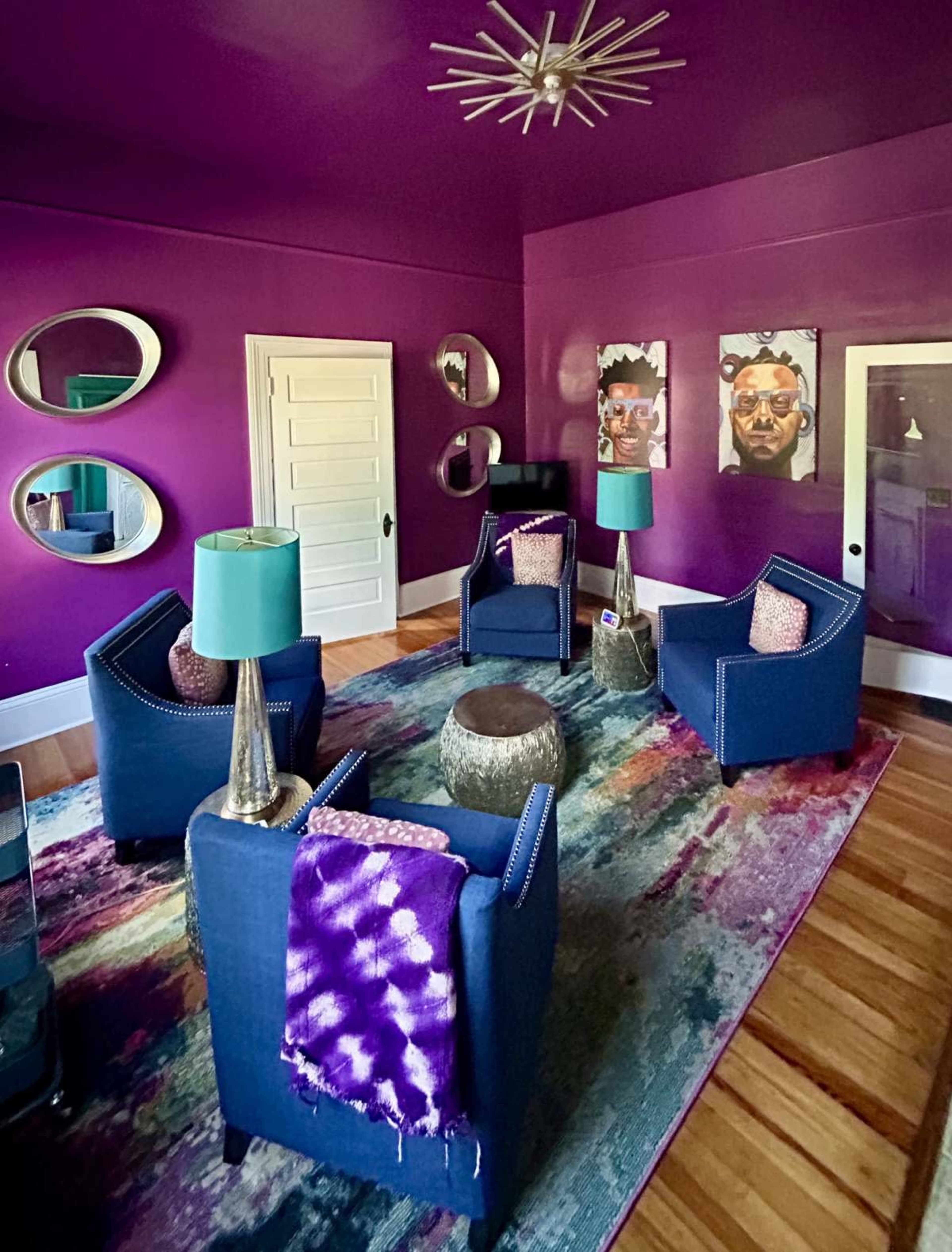A purple-walled room features two blue chairs with decorative cushions, a central table, and a colorful rug, accompanied by mirrors and artwork on the walls.
