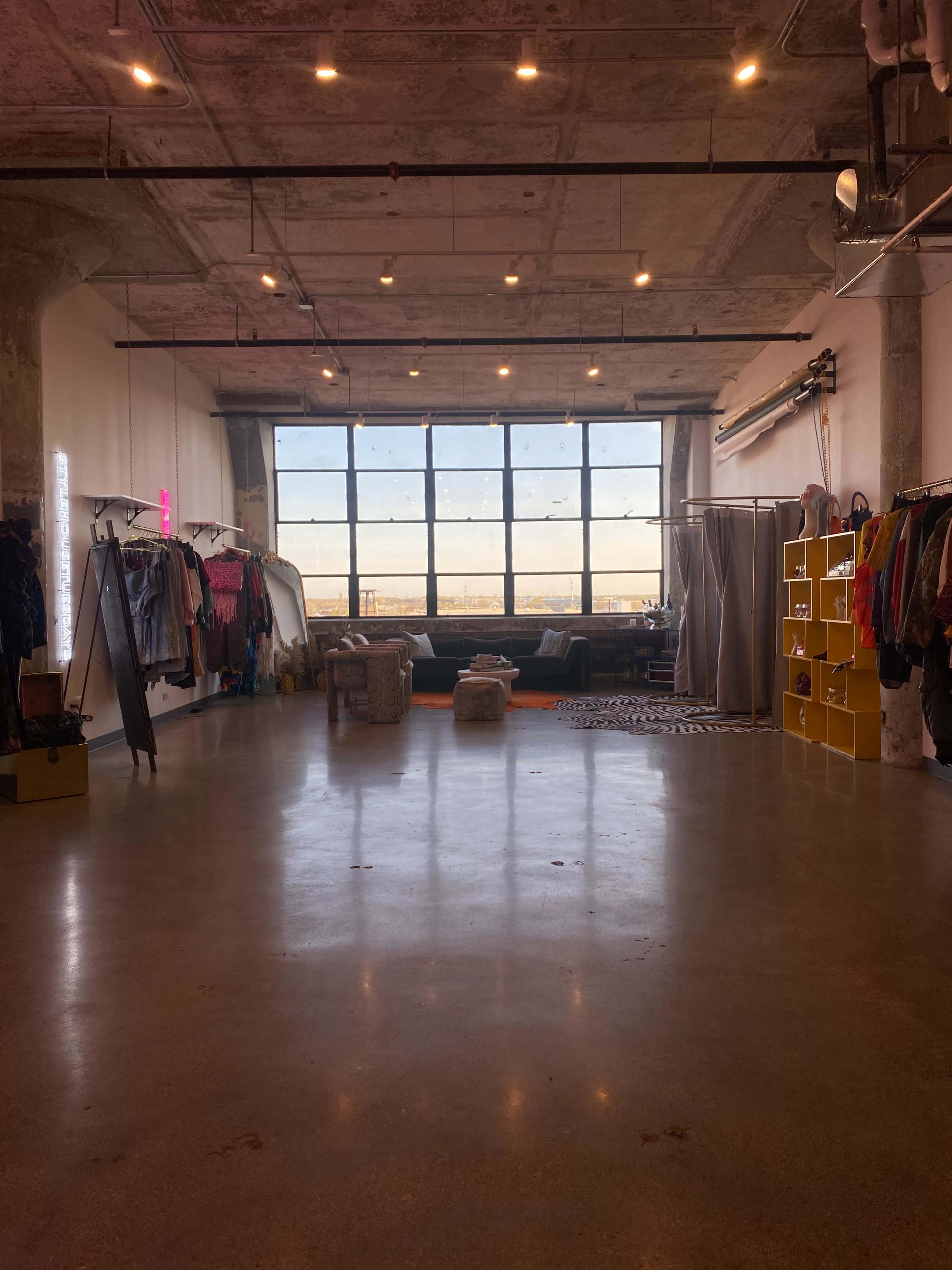 The image shows an industrial-style room with a large window, clothing racks along the walls, and a seating area in the background.