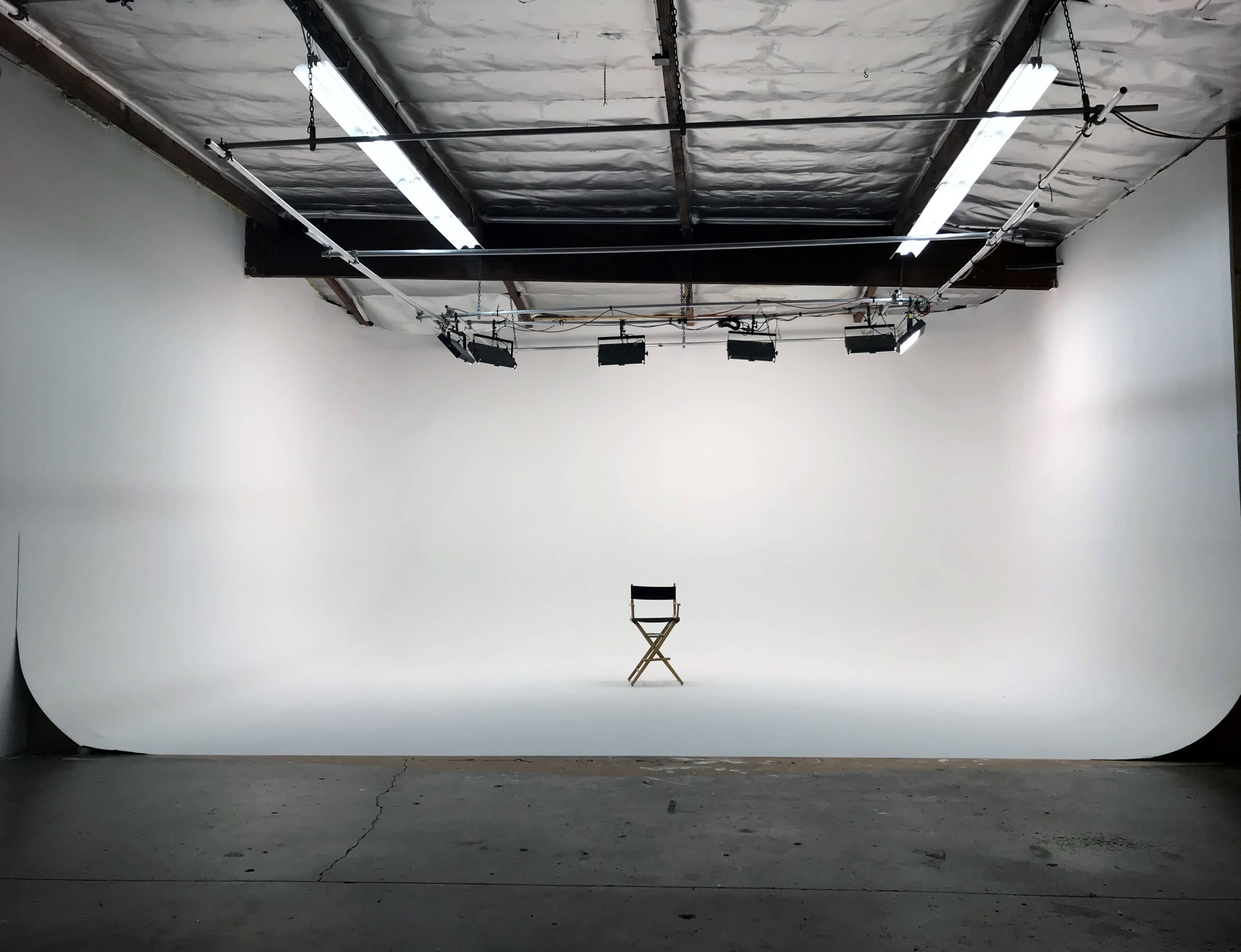 An empty studio space with a wooden director's chair positioned on a seamless white background under overhead lights.