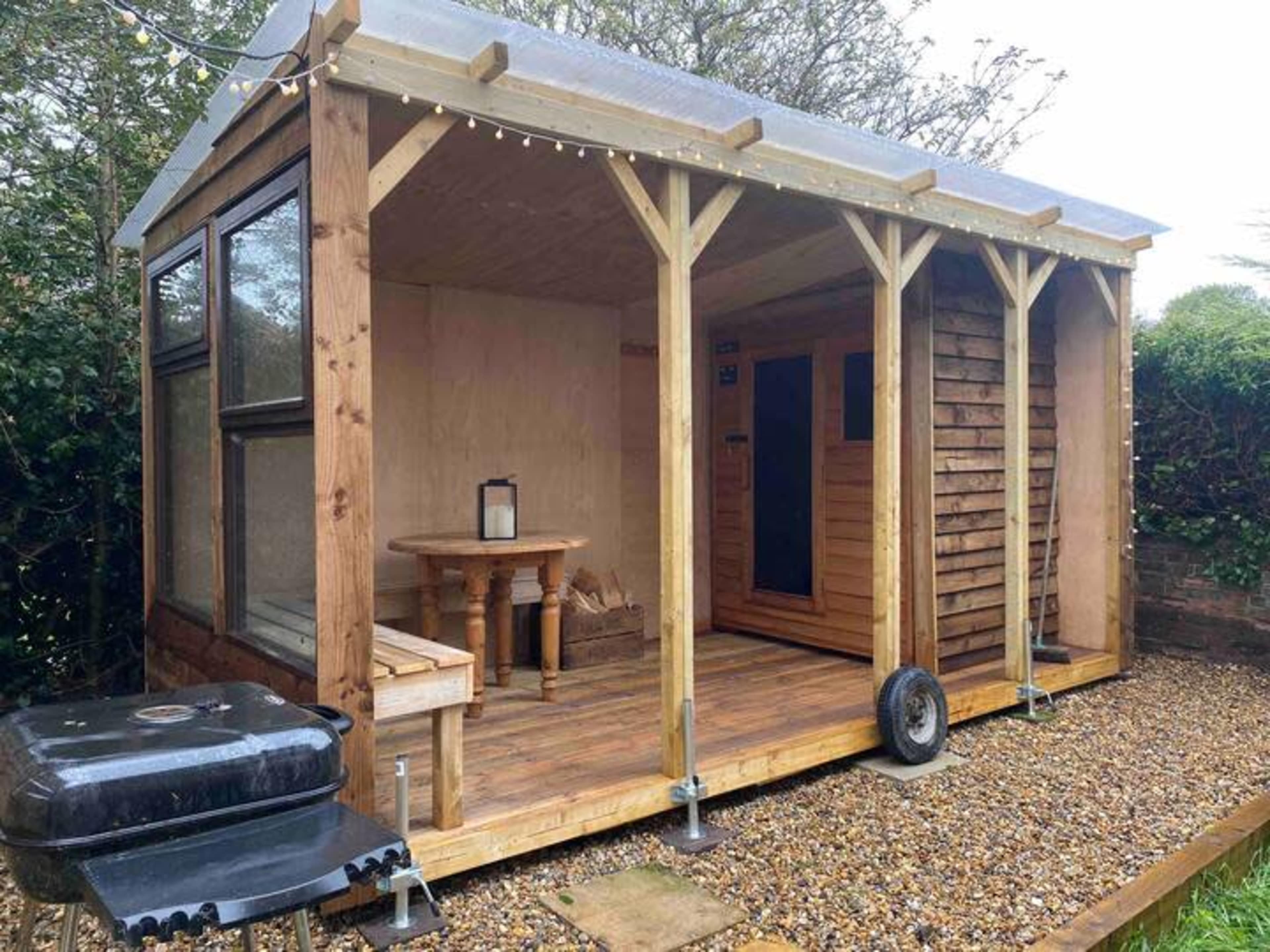 The image shows a wooden garden shed with a porch, featuring a table, a black grill, and a sauna inside.