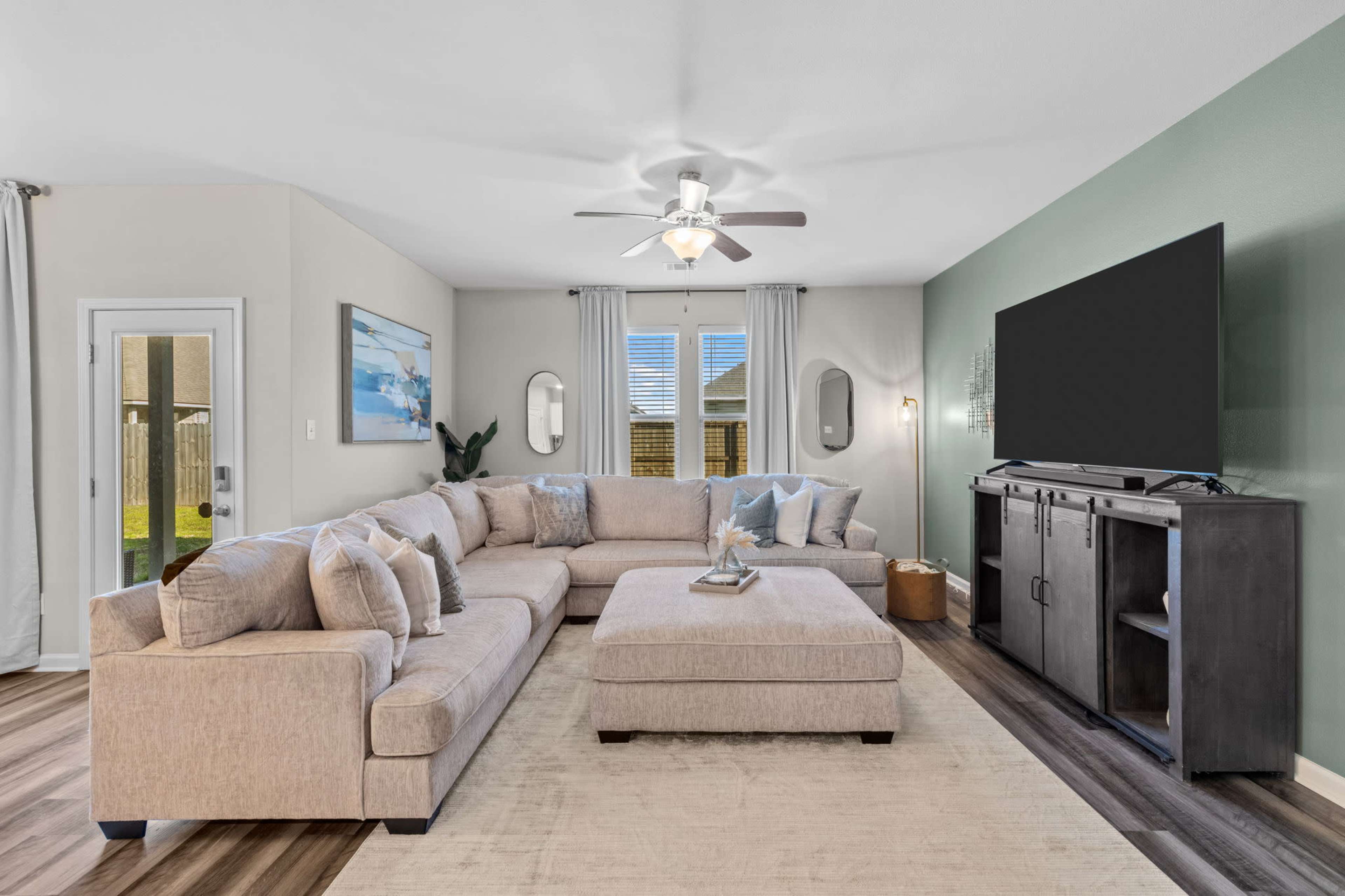 A spacious living room with a large sectional sofa, a television mounted on the wall, and light-colored decor.