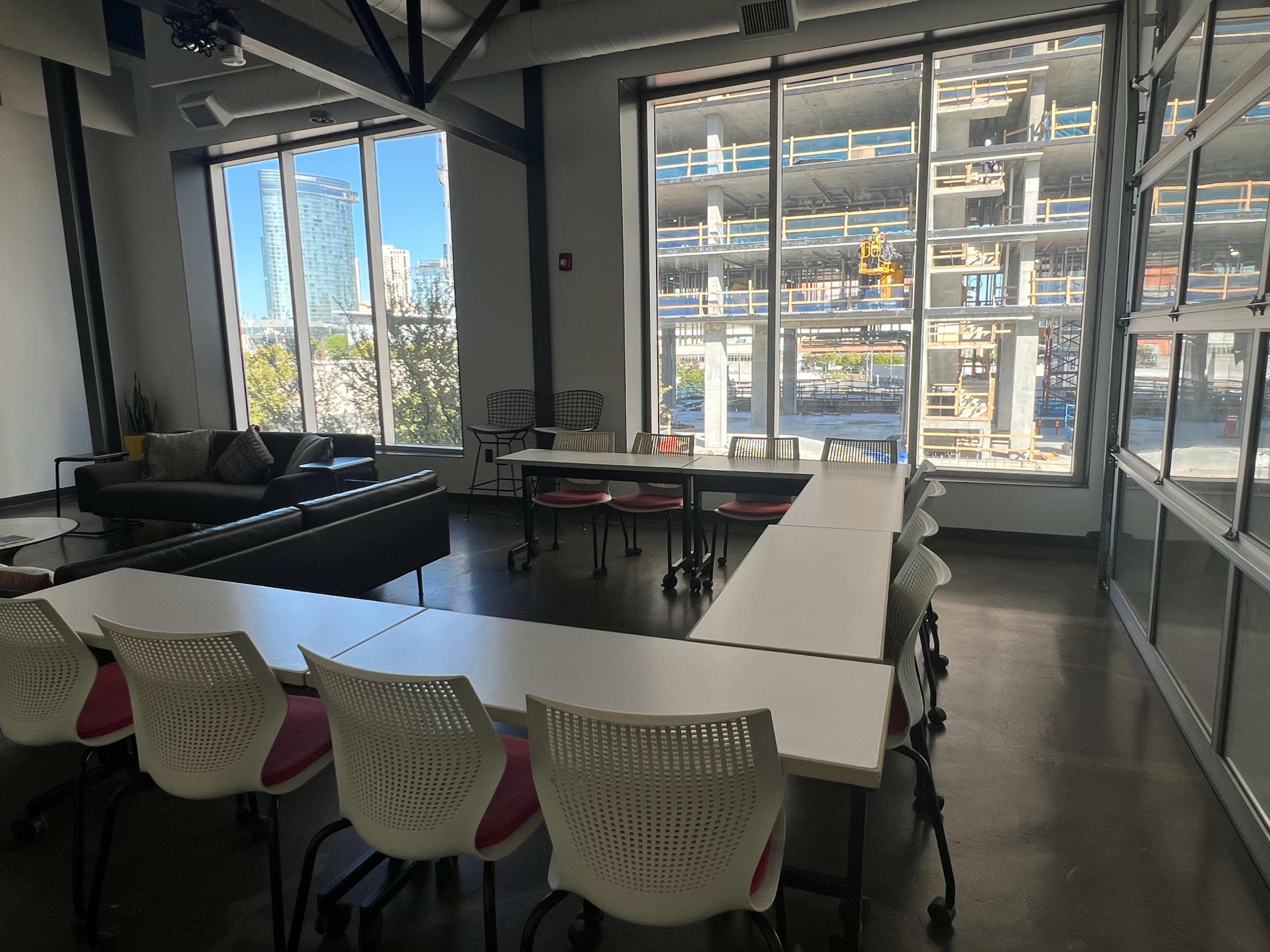 A modern conference room features a long white table surrounded by ergonomic chairs, with large windows overlooking a construction site and city skyline.