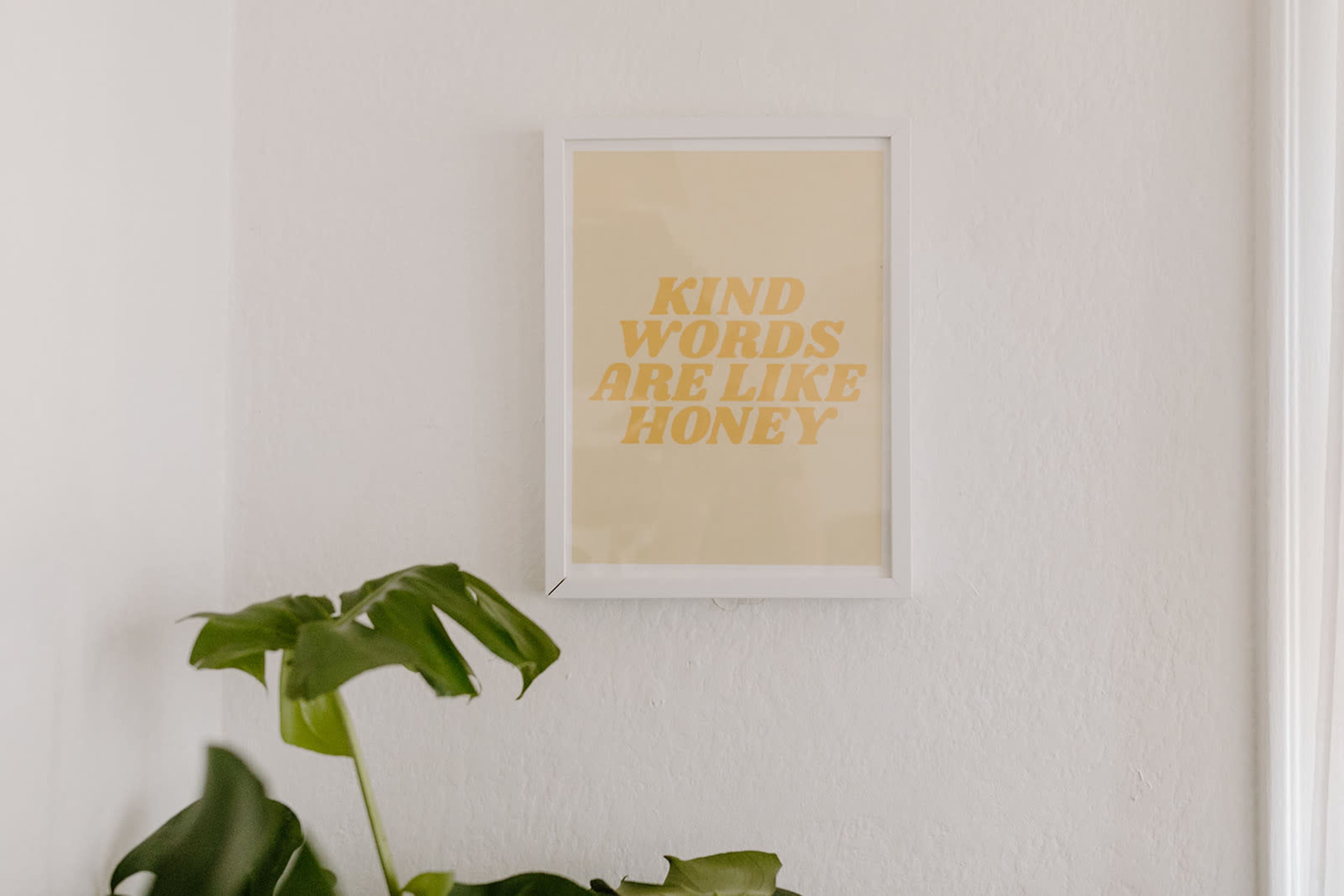 A framed poster with the text "Kind words are like honey" hangs on a white wall next to a green plant.