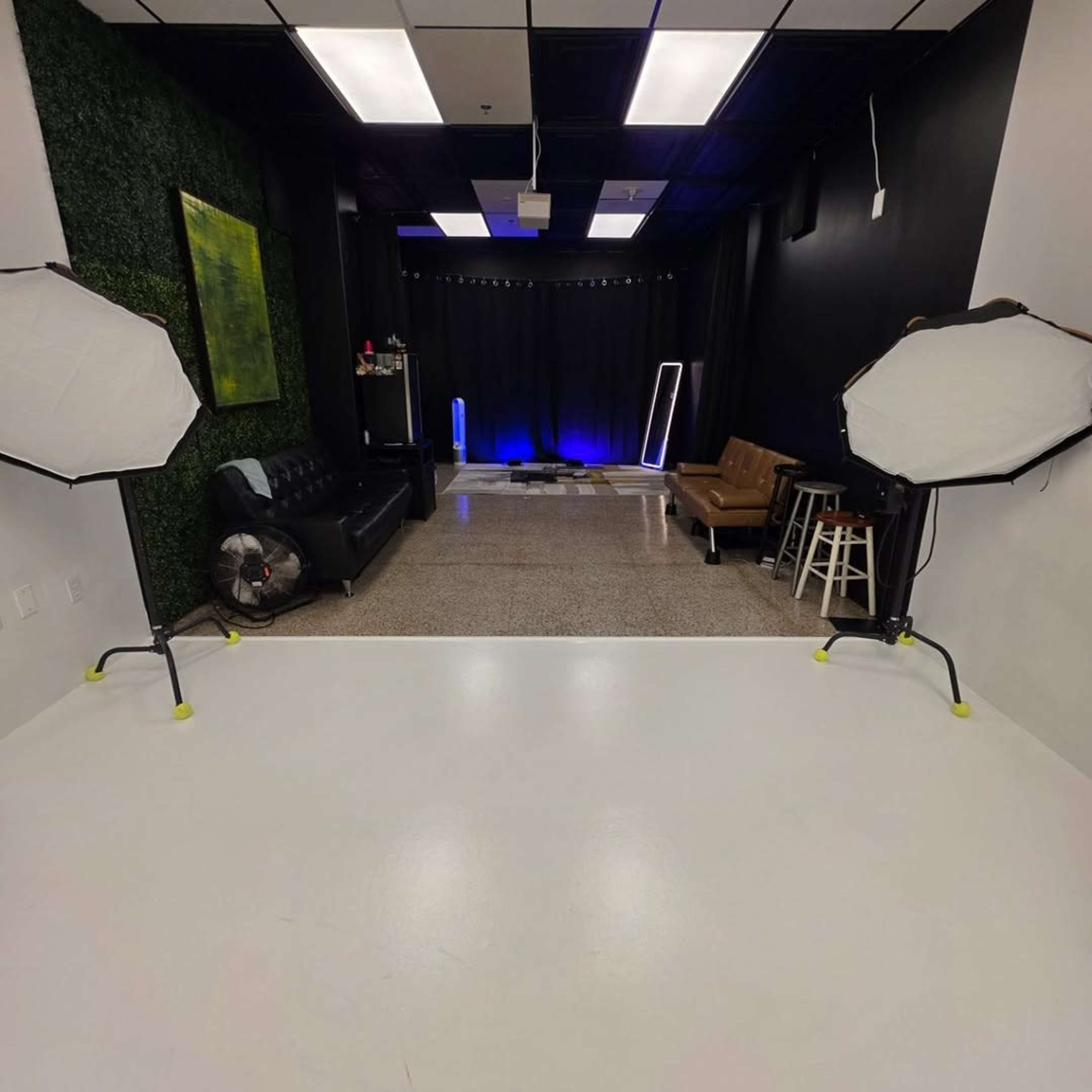 The image shows a photography studio with two softbox lighting setups, a black backdrop, and various furniture including a leather couch and stools.