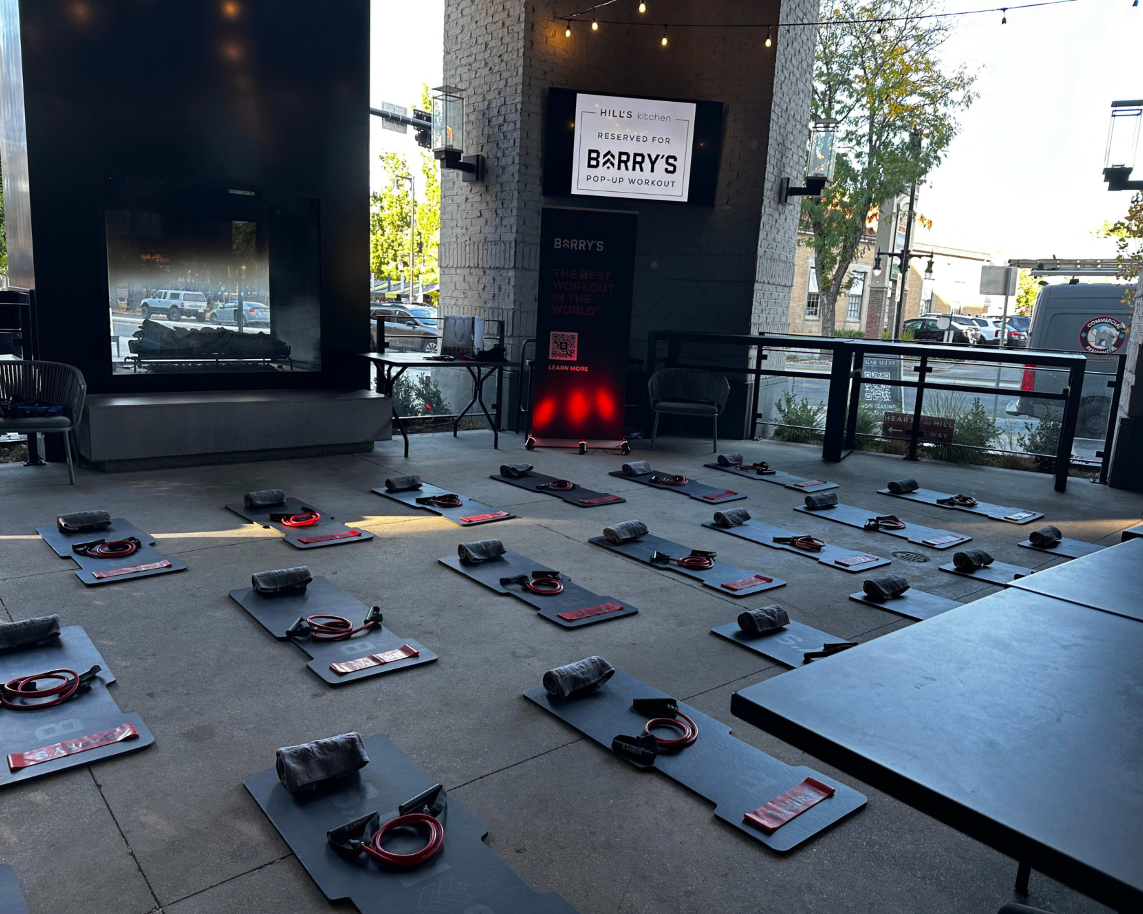 The image shows a fitness studio setup with several workout mats arranged in rows on a patio, each equipped with red resistance bands.