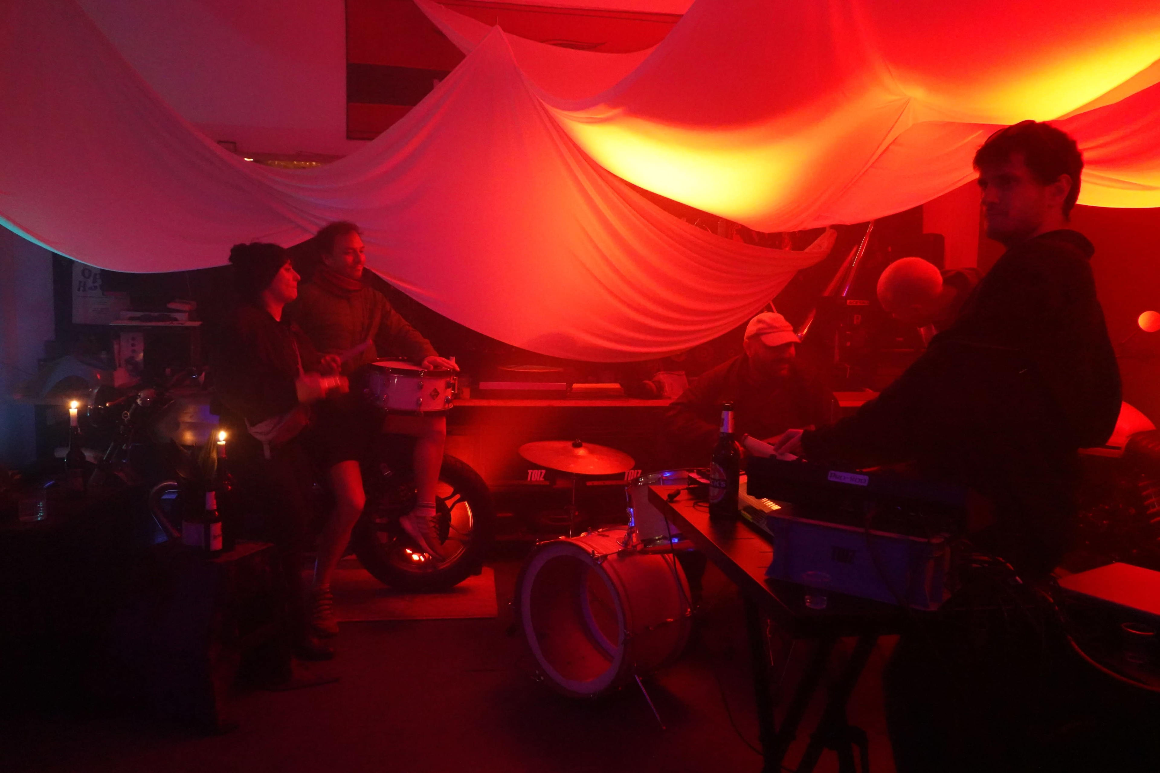 A group of musicians is playing instruments and socializing in a dimly lit room adorned with draped fabric and ambient lighting.