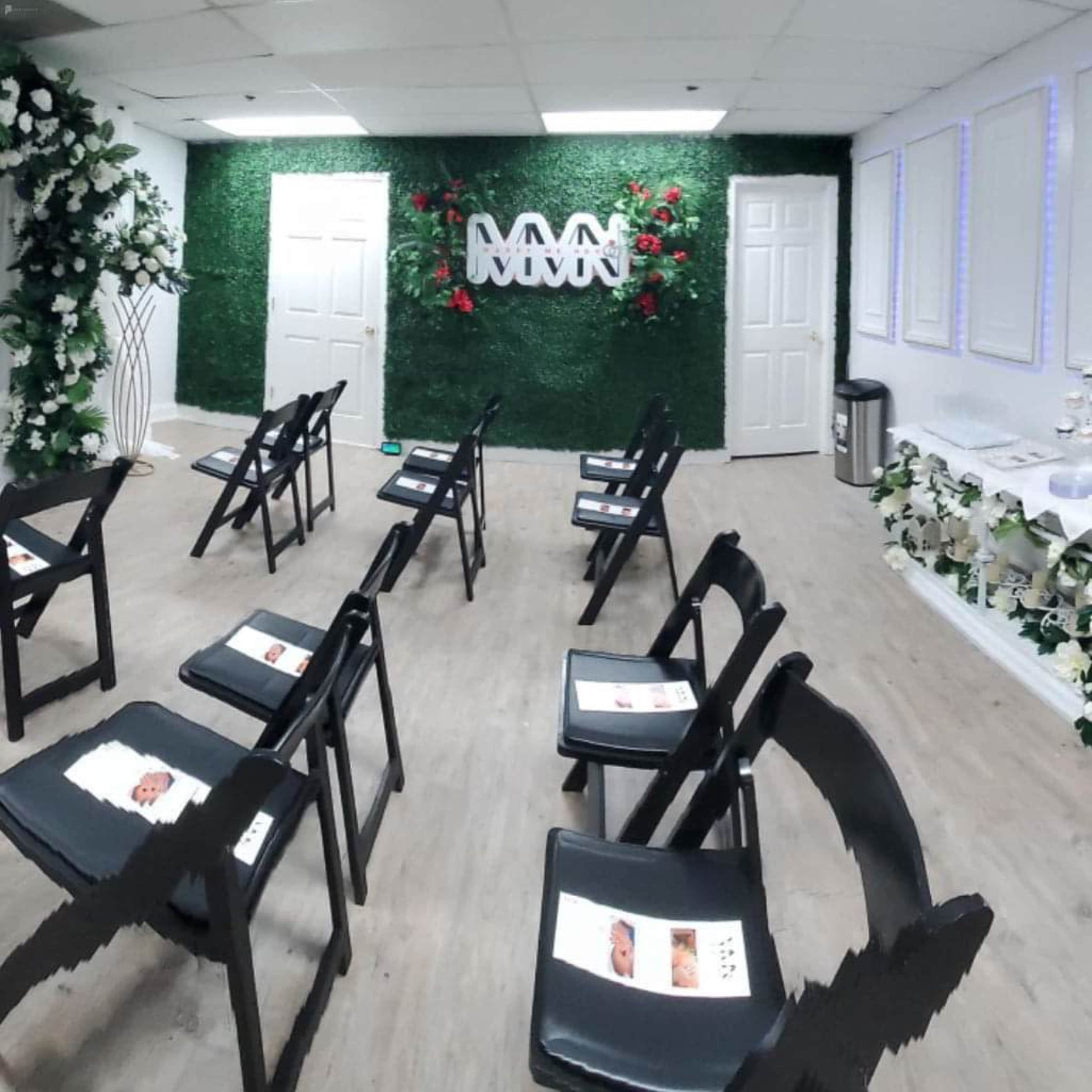 A room with black folding chairs arranged in front of a green wall decorated with plants and flowers, featuring three white letters on the wall.