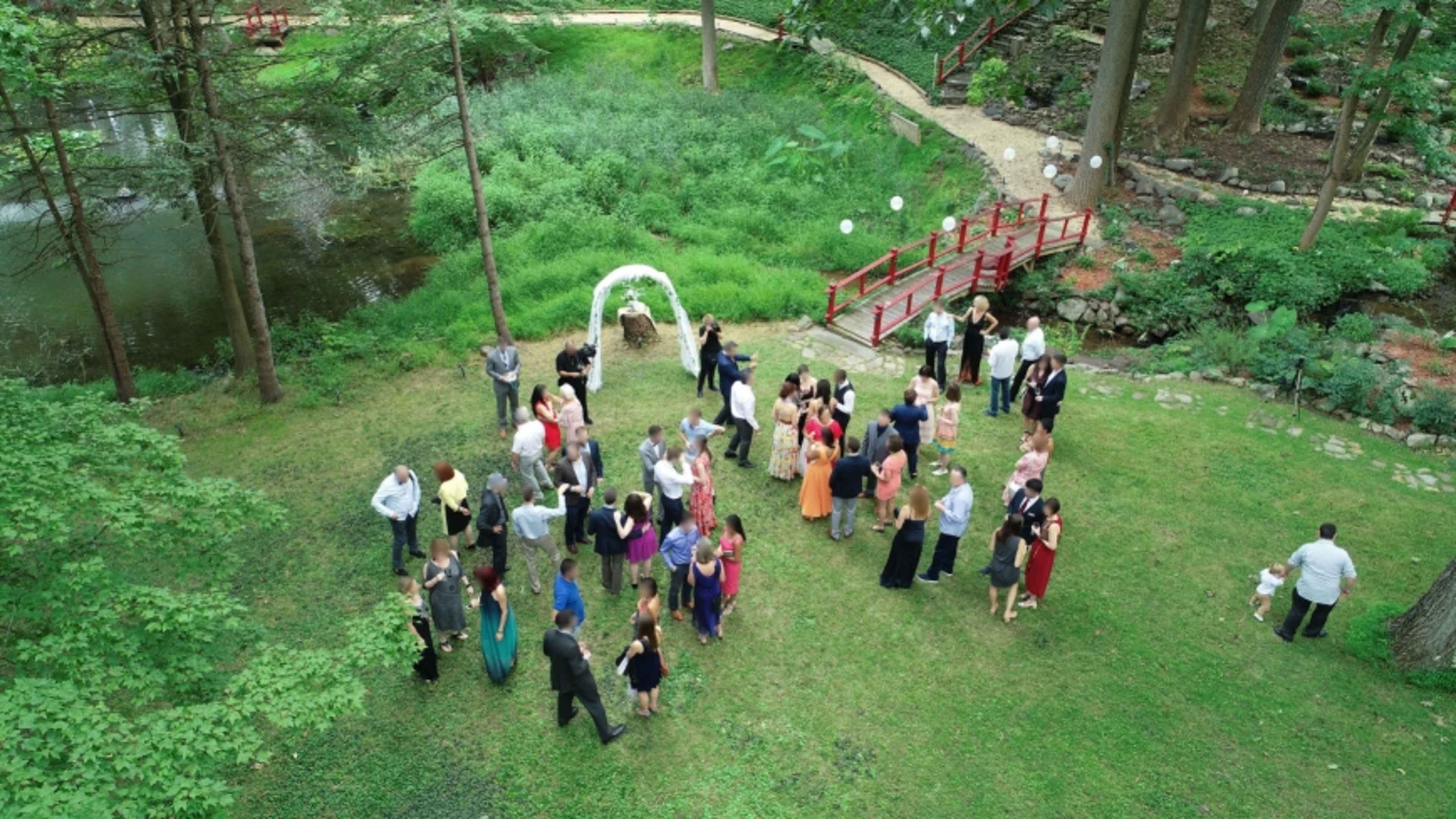 A large outdoor gathering is taking place on a grassy area near a pond, with guests mingling around an arch and a red bridge in the background.