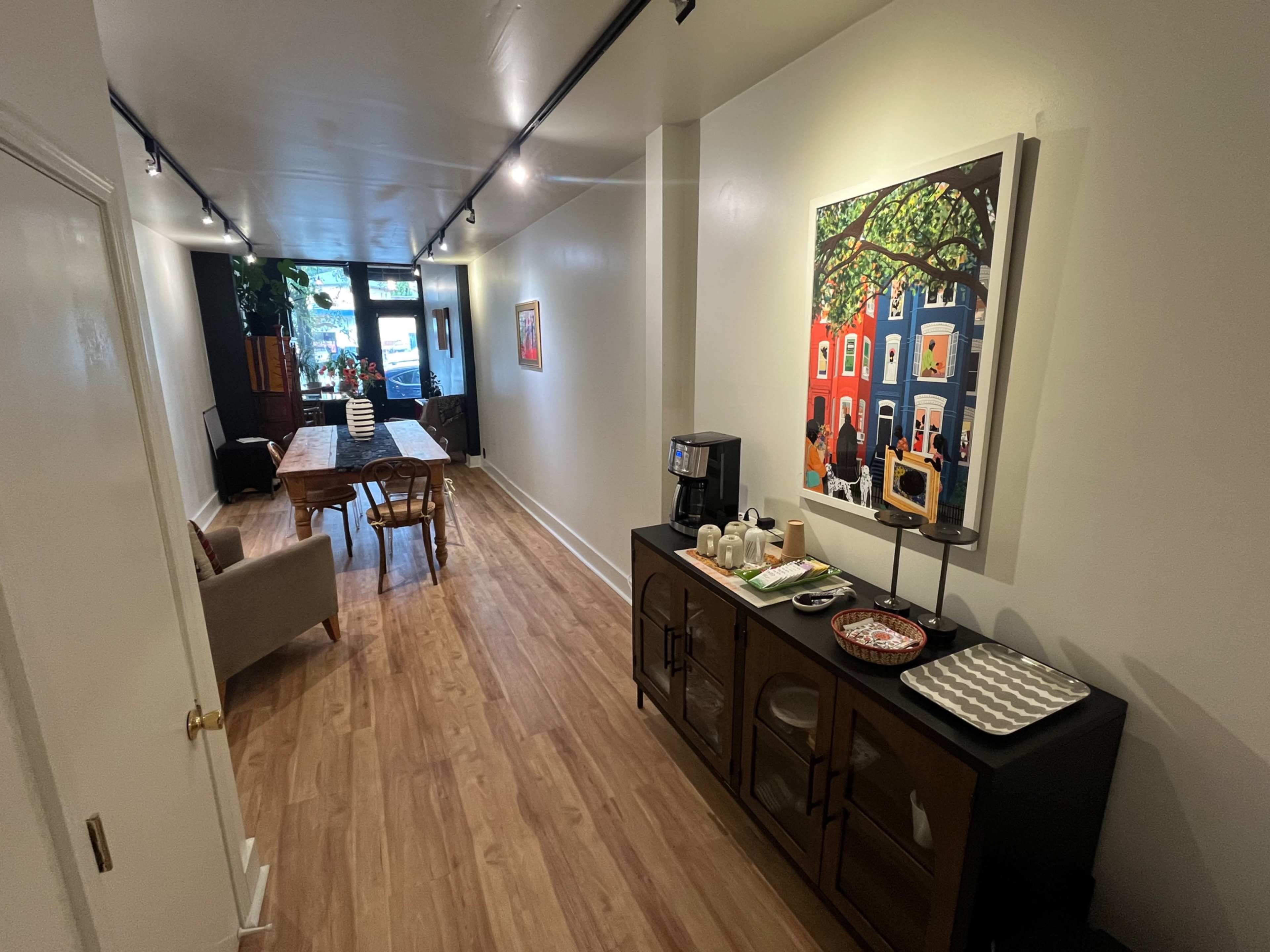 A long, narrow room features a dining table, seating area, and artwork on the wall, with natural light streaming in from a large front window.