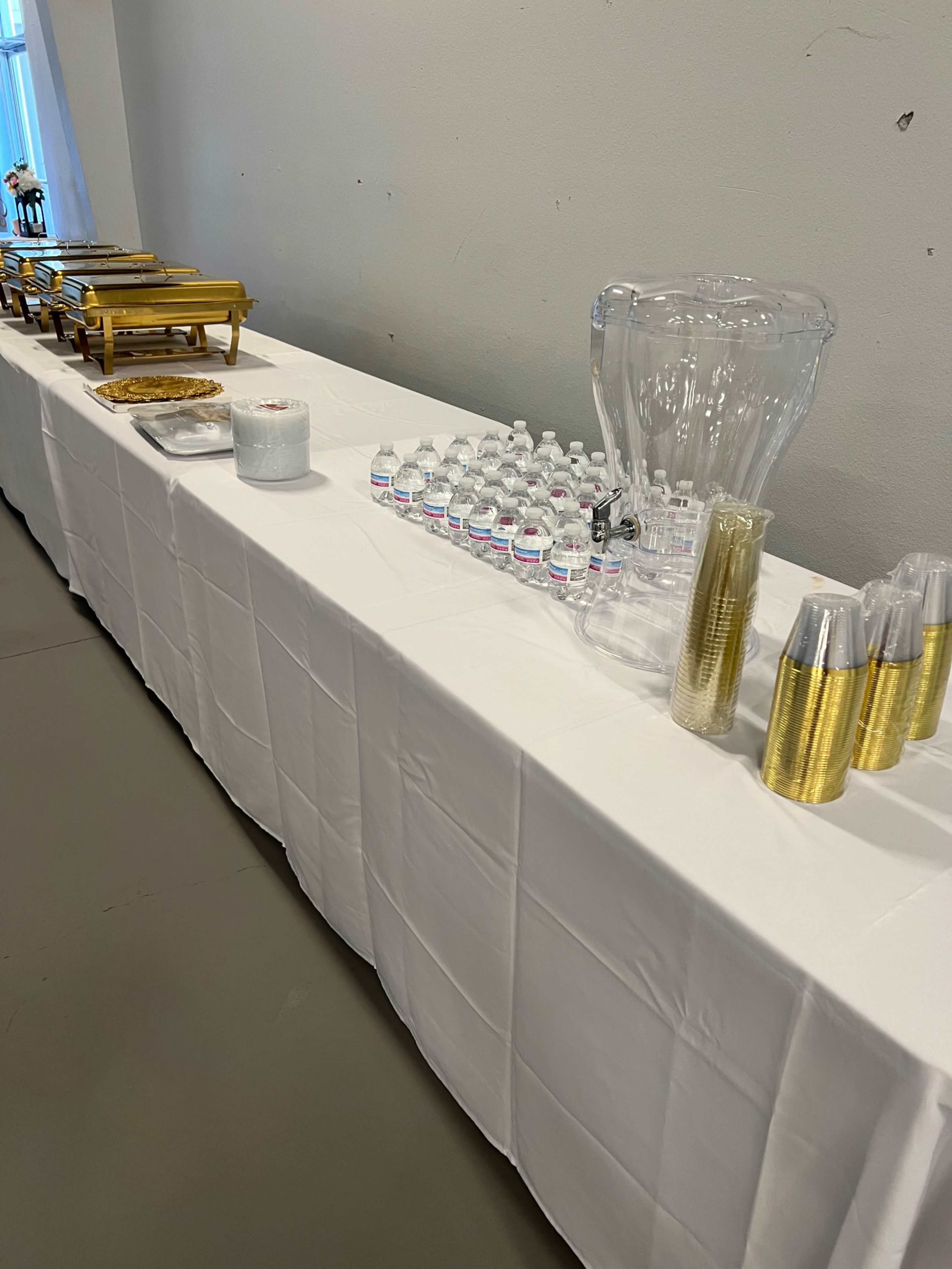 The image shows a table covered with a white tablecloth, featuring chafing dishes, a water dispenser, and an arrangement of plastic cups and bottled water.