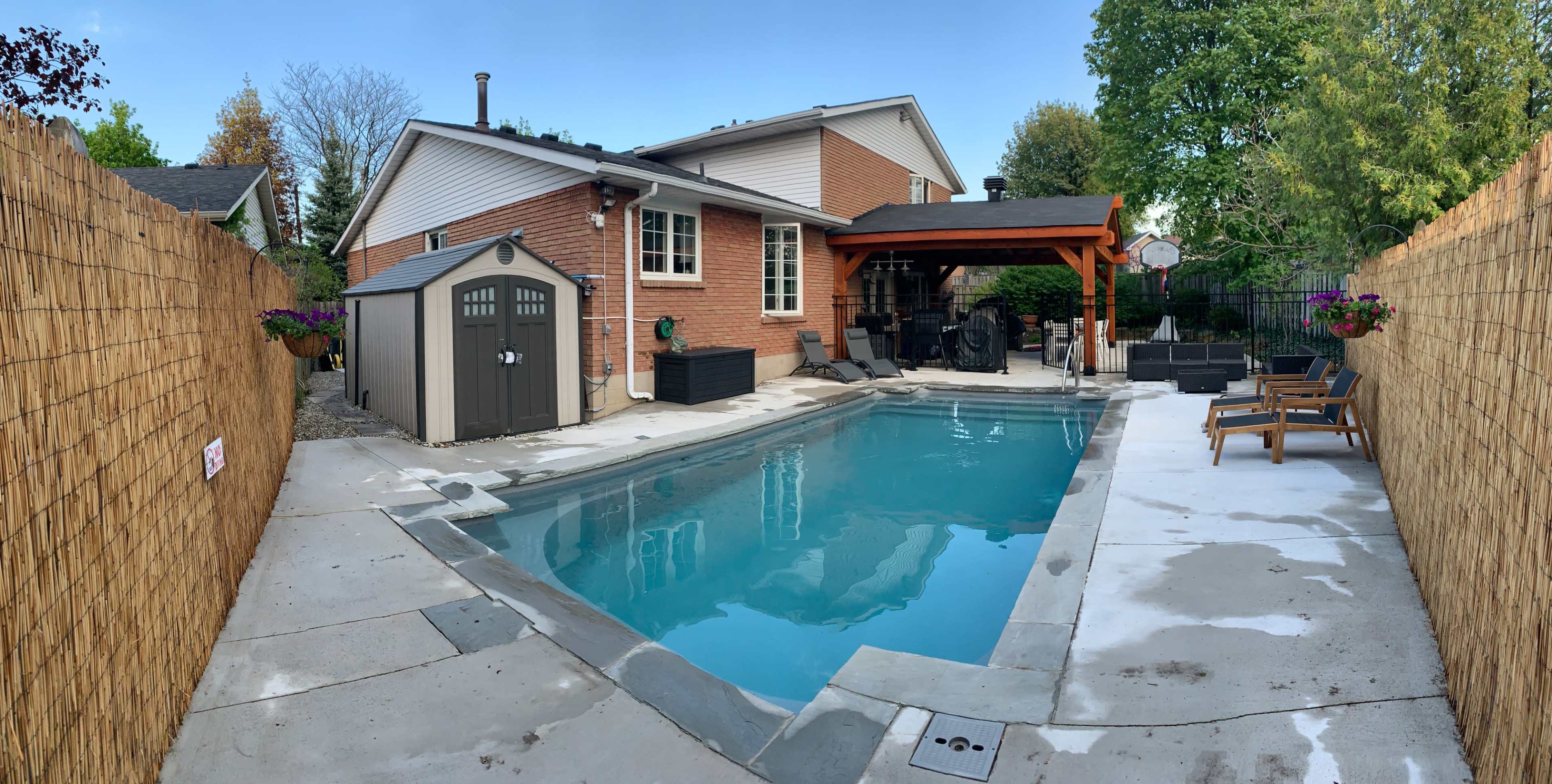 The image shows a residential backyard featuring a swimming pool surrounded by a concrete patio, a storage shed, and outdoor seating under a covered area.