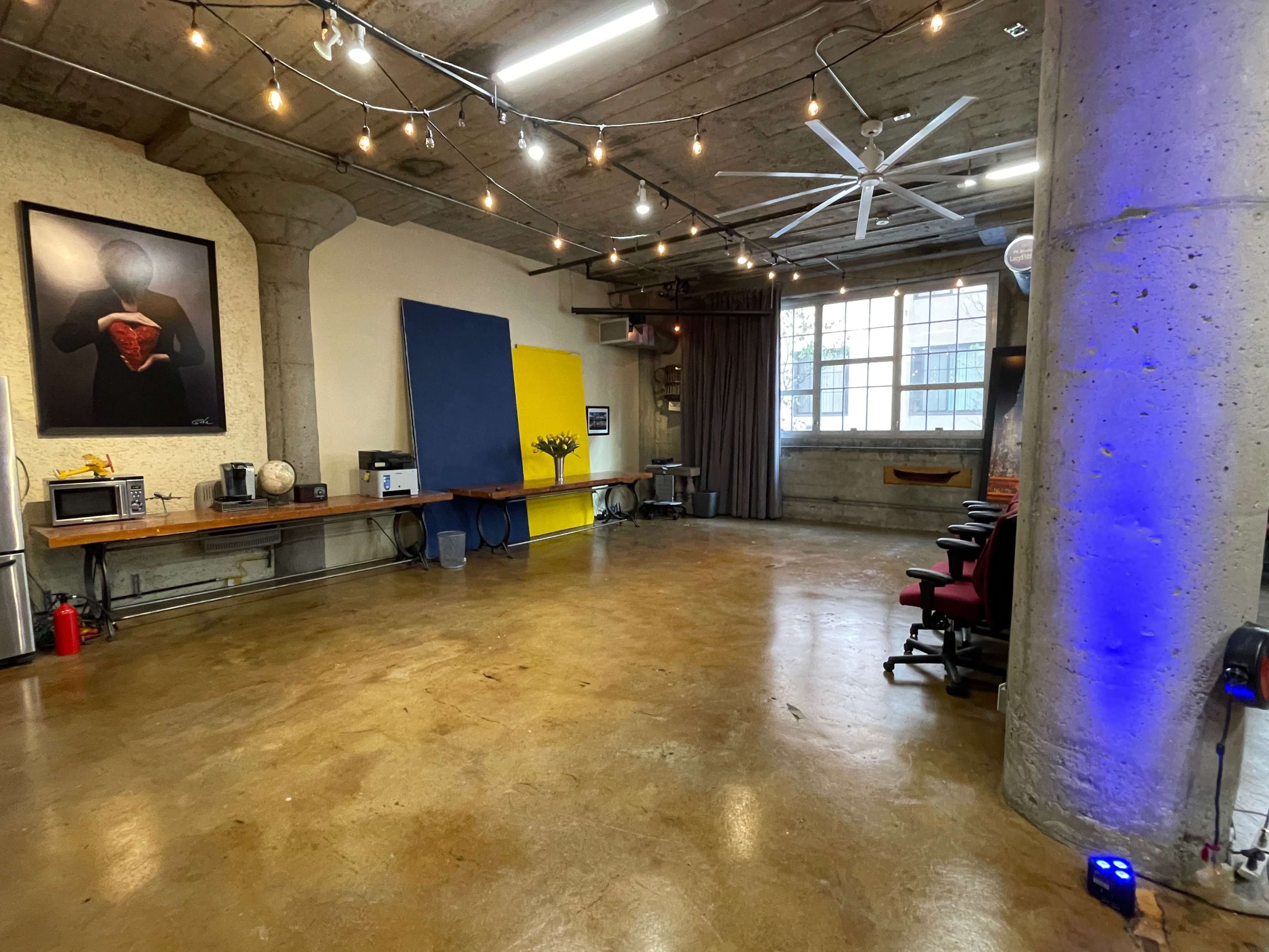 A spacious studio with concrete walls, wooden tables, a large bright blue canvas, and string lights overhead.