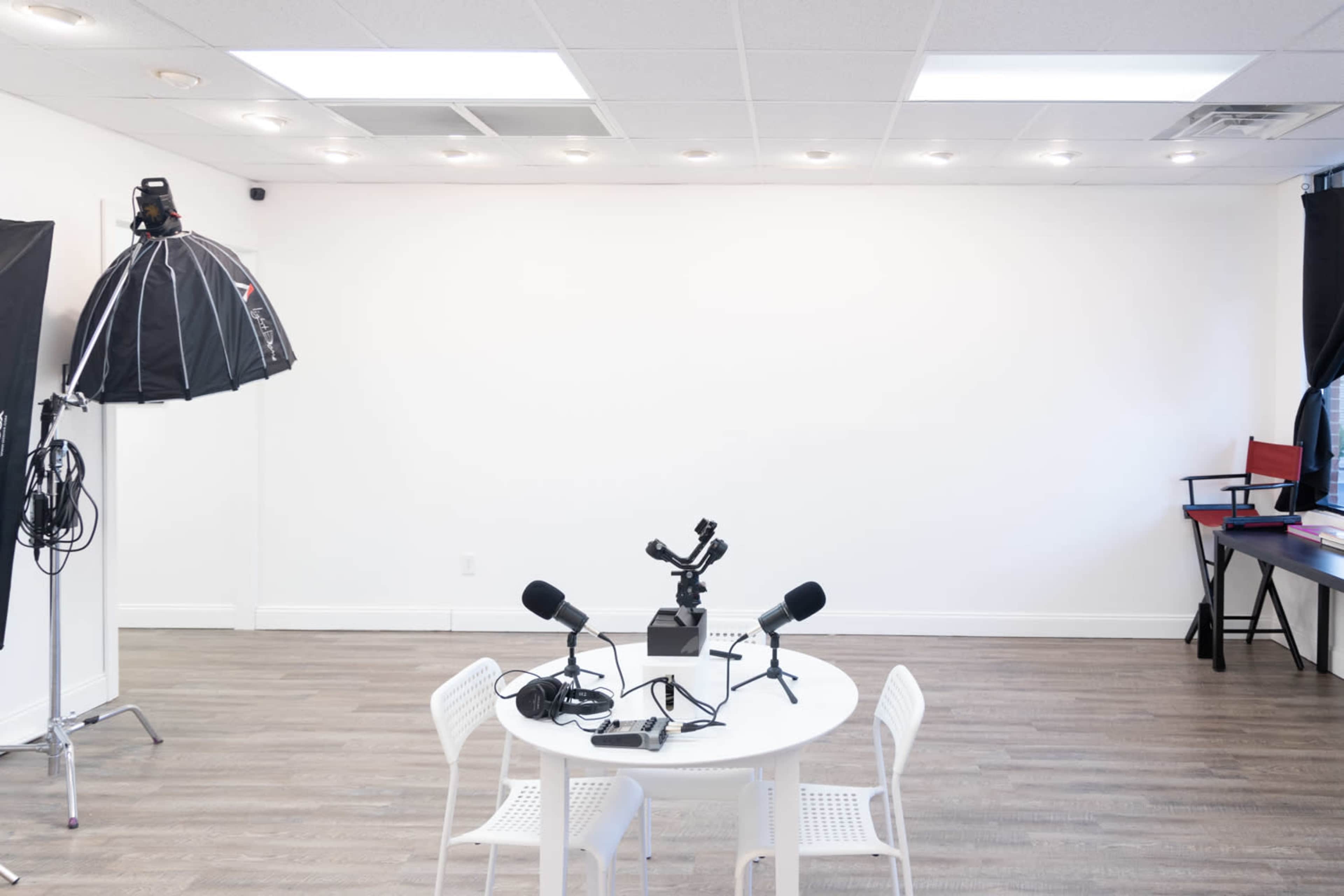 A small, empty studio space features a round table with microphones and recording equipment, surrounded by white walls and wooden flooring.