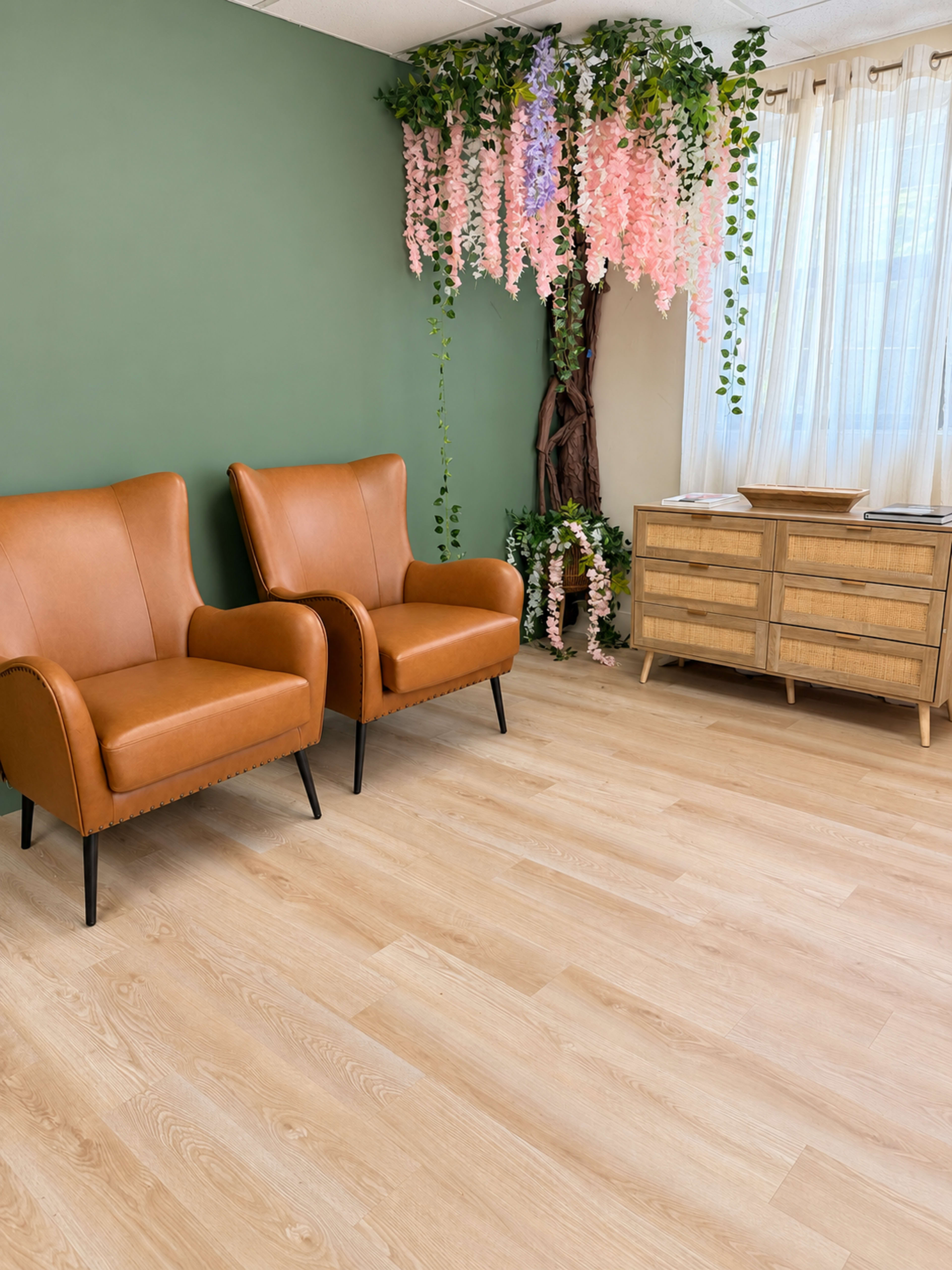 The image features a cozy interior space with two brown armchairs facing each other, a wooden dresser, and a lush floral arrangement hanging from the ceiling.