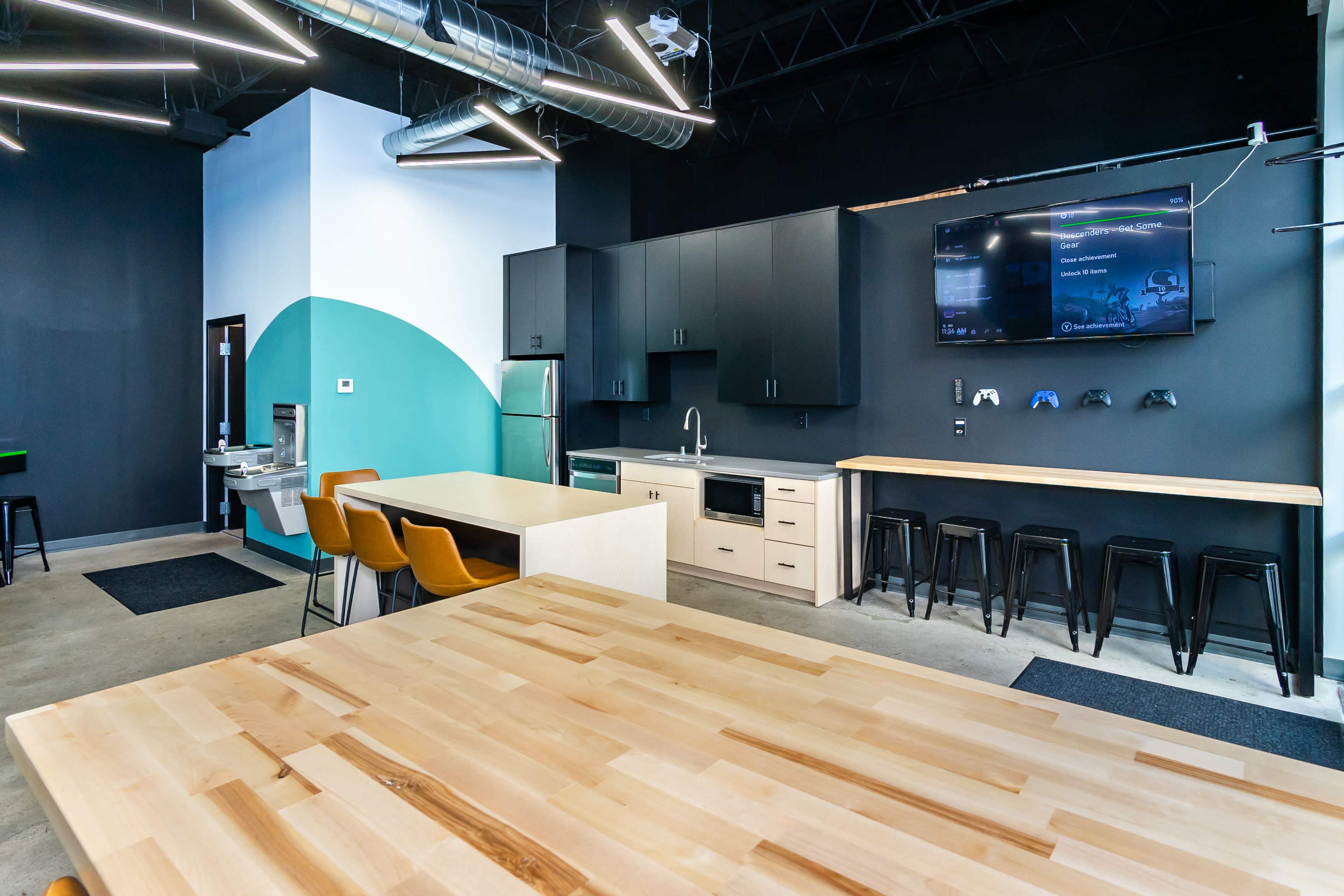 The image shows a modern kitchen and dining area with a large wooden table, sleek black cabinetry, and a wall-mounted TV displaying content.