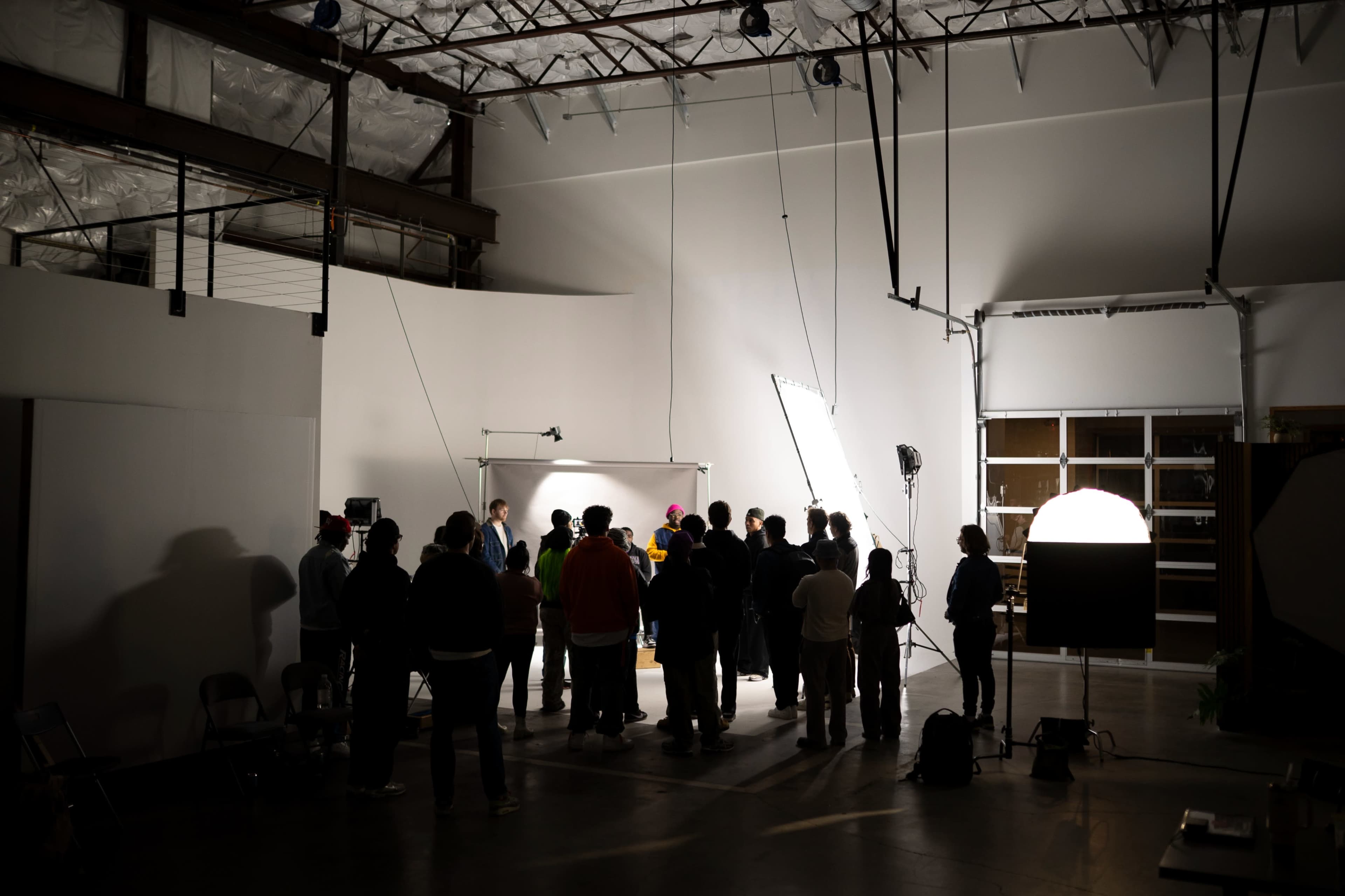 A group of people is gathered in a well-lit studio space, focusing on a subject illuminated by multiple light sources.