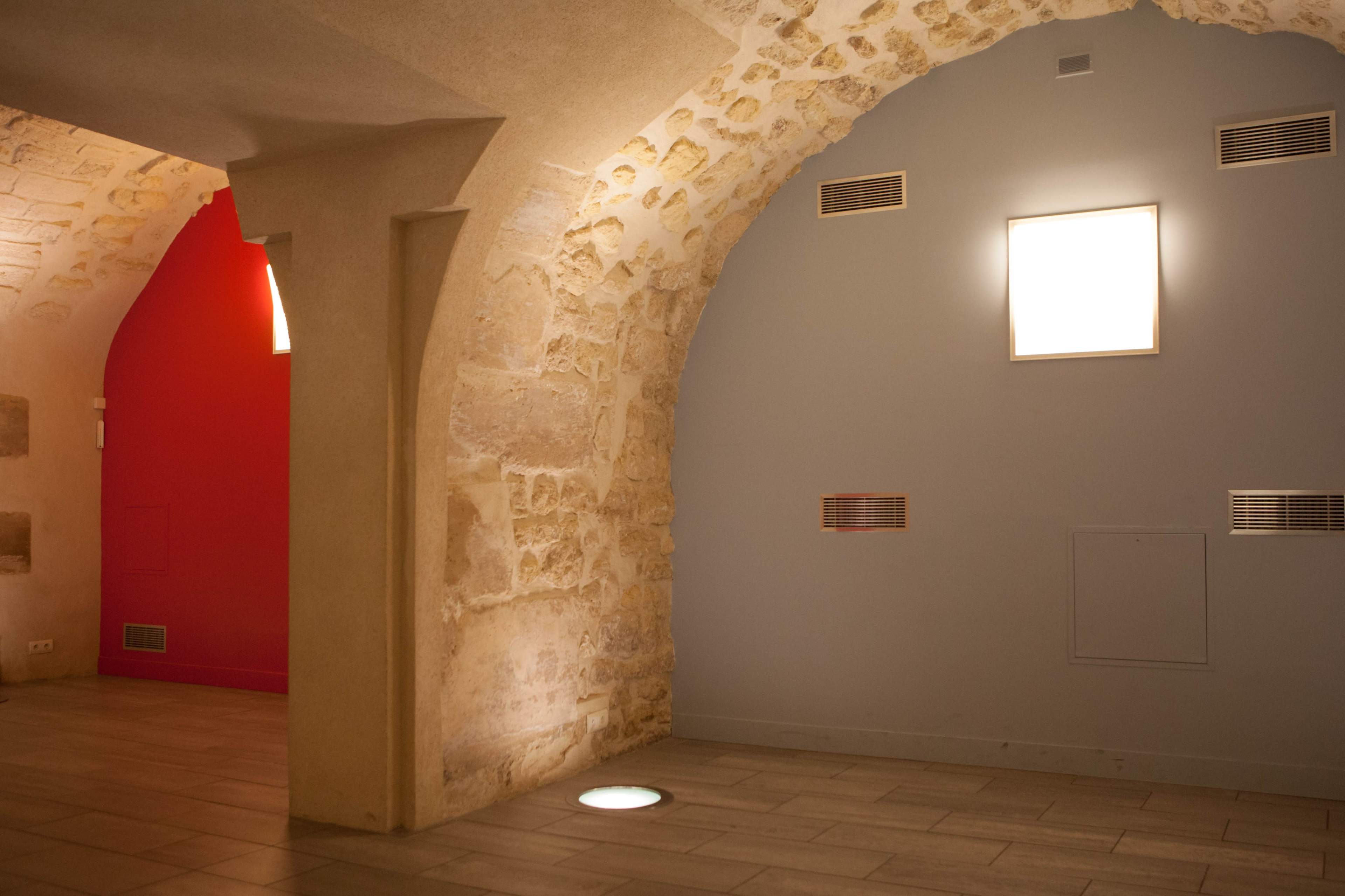 The image shows a stone-walled interior space with two arched sections, one featuring a red wall and the other painted gray, illuminated by square light fixtures.