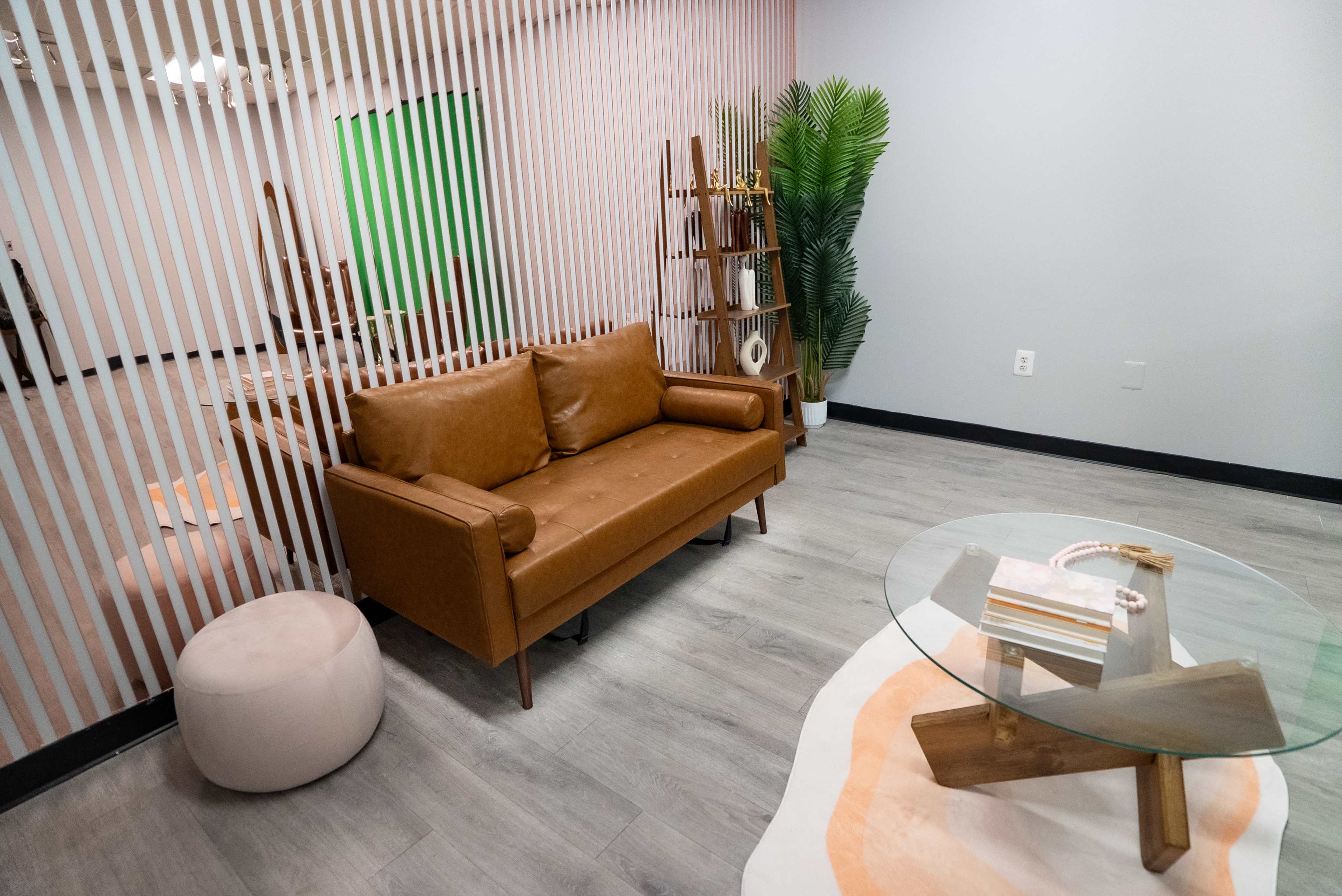 The image shows a modern lounge area with a brown leather couch, a glass coffee table, and a soft pouf, separated by vertical slatted partitions from an open space.