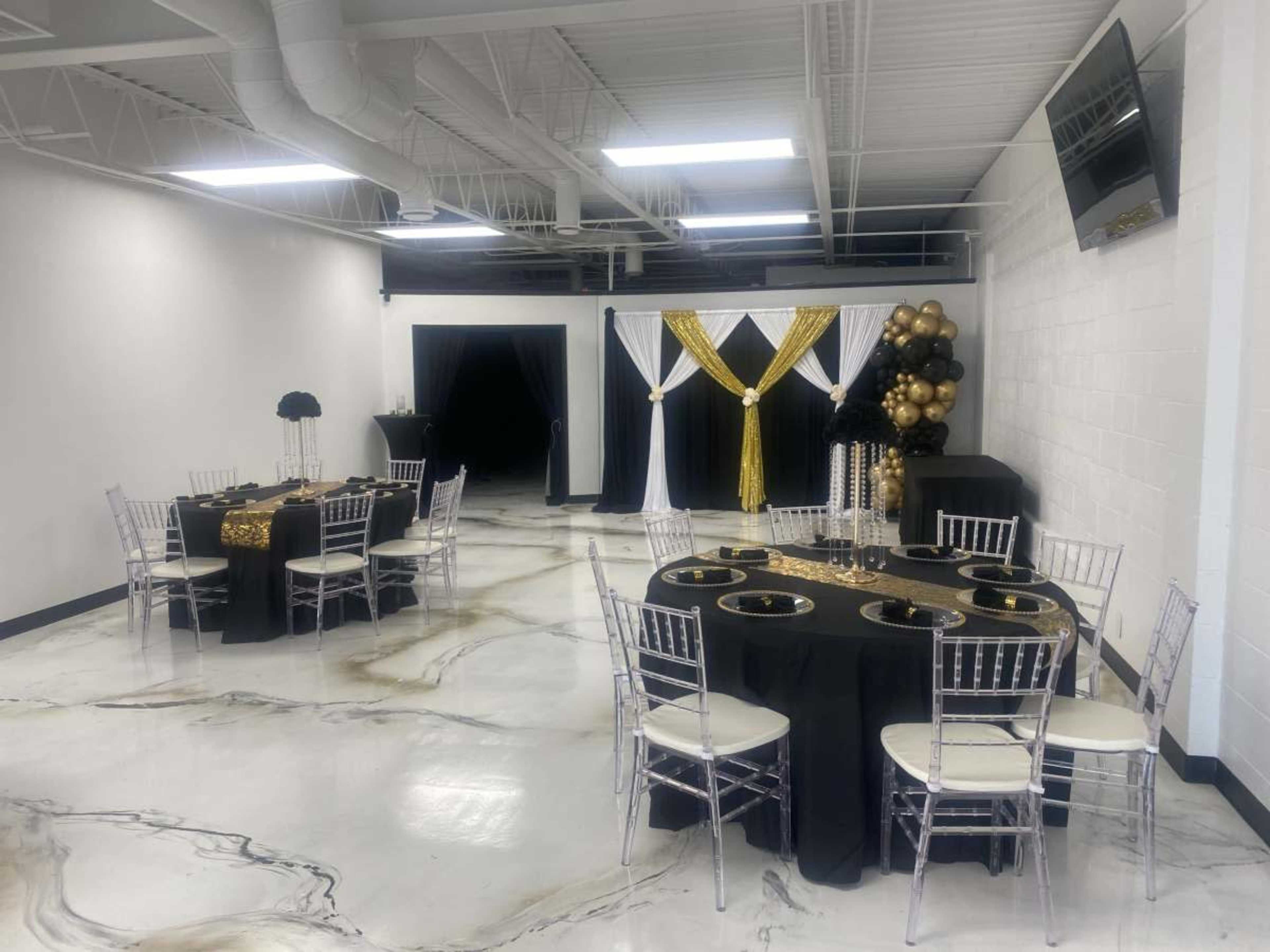 A banquet hall features black and gold decorated tables set for an event, with elegant chairs and a backdrop draped in white and gold fabric.