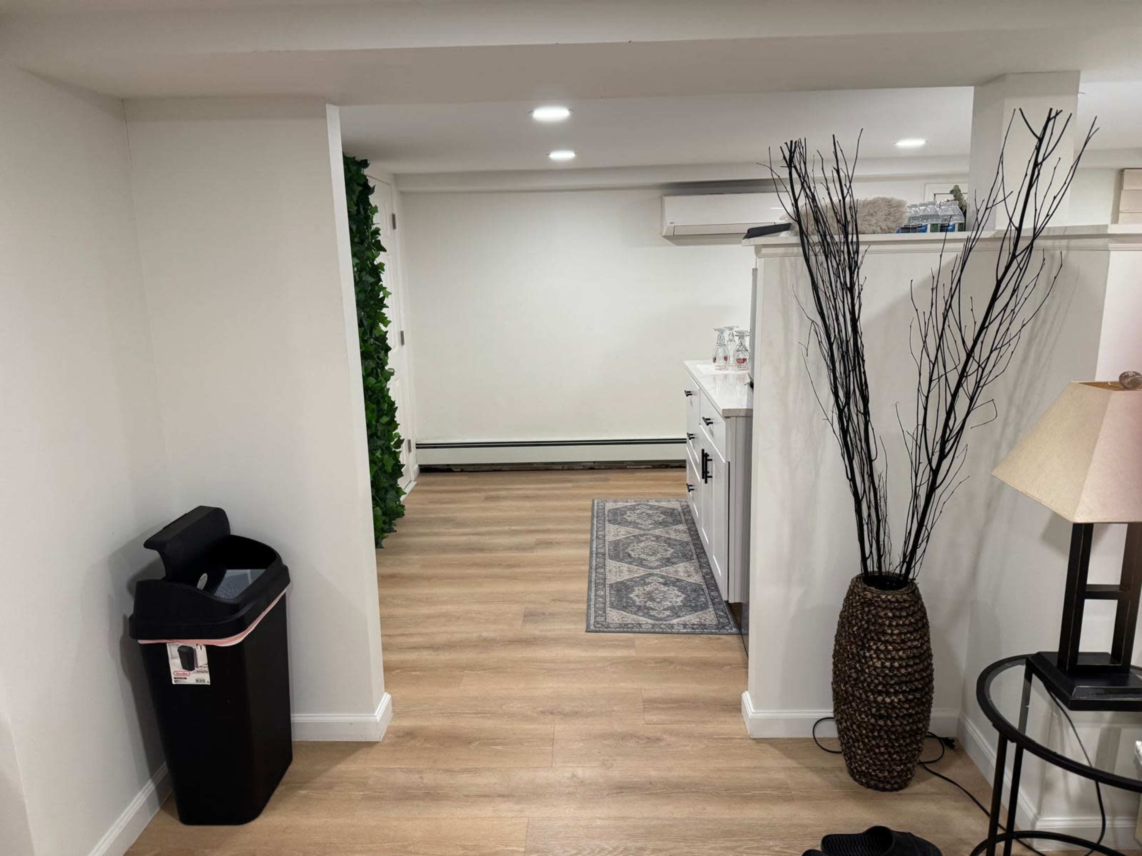 The image shows a well-lit interior space with wooden flooring, featuring a trash bin, a decorative plant, and furniture including a lamp and a side table.
