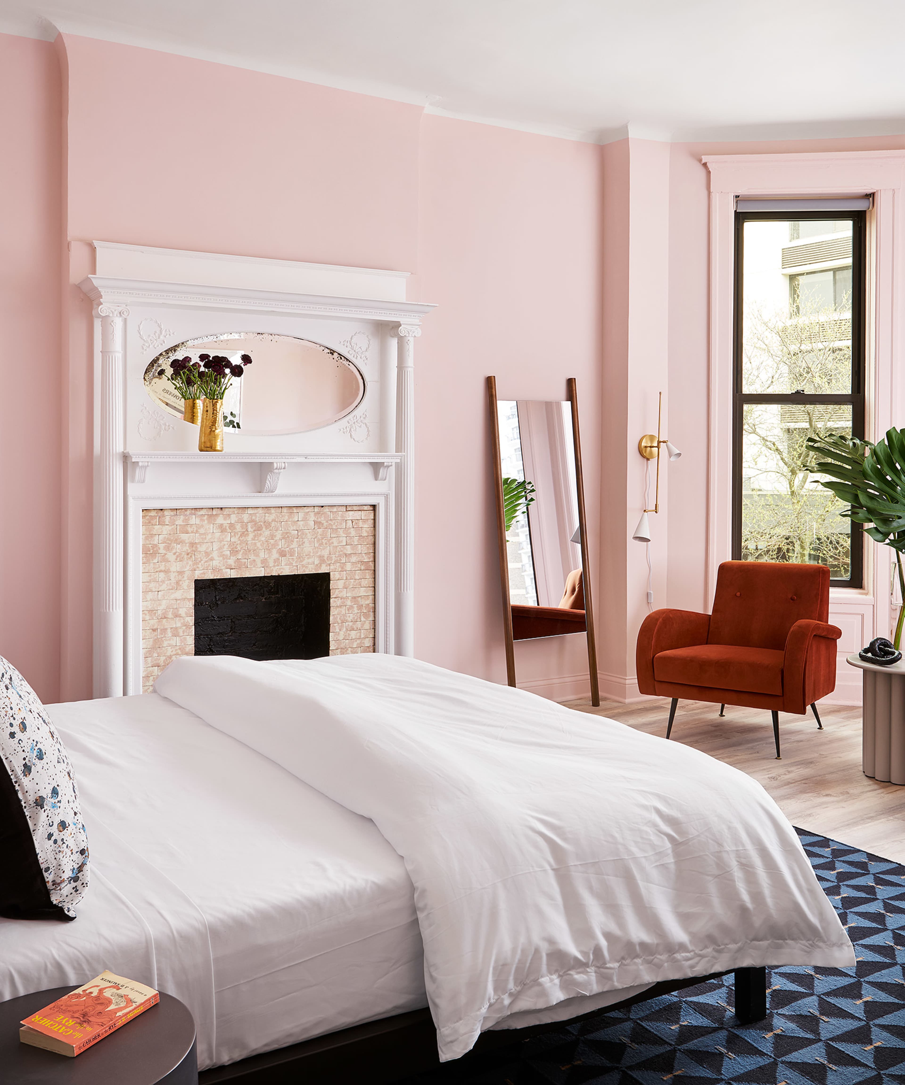 The image shows a bedroom with a light pink wall, a white bed covered with a duvet, a fireplace with a mirror above it, a standing mirror, an orange armchair, and a potted plant.