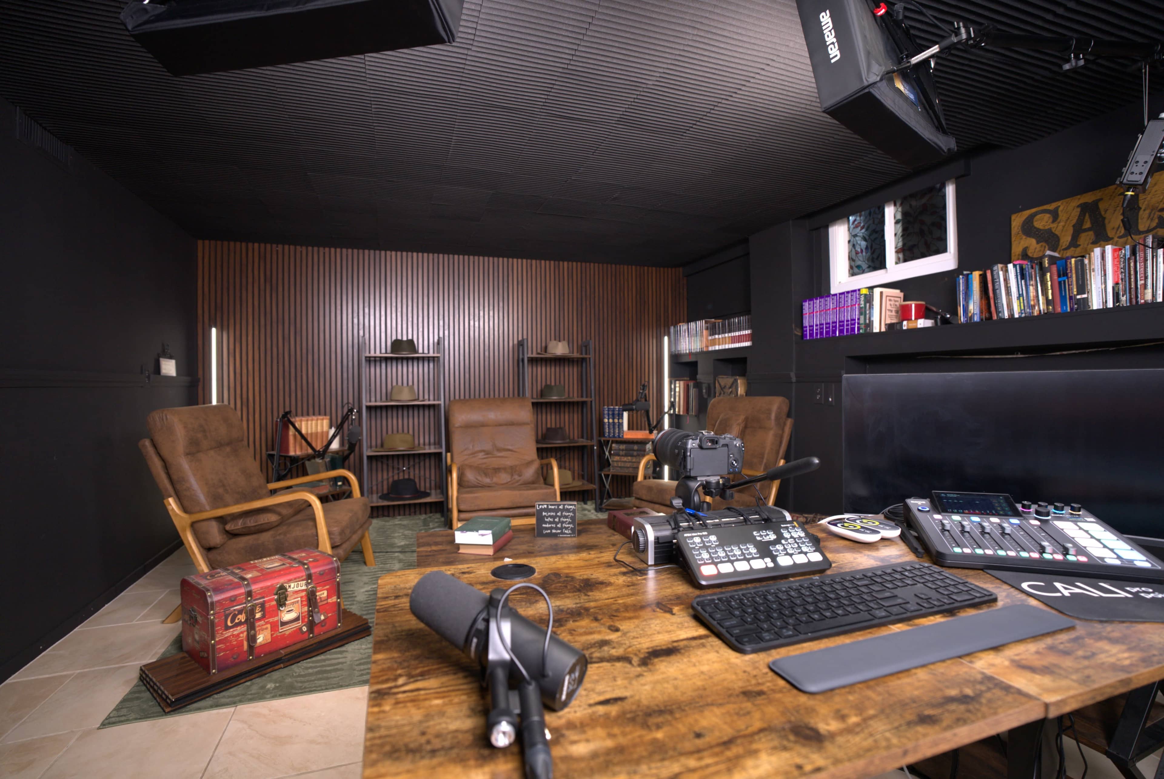 The image depicts a modern recording studio with two armchairs, shelves filled with books, and a desk equipped with audio equipment and a camera.