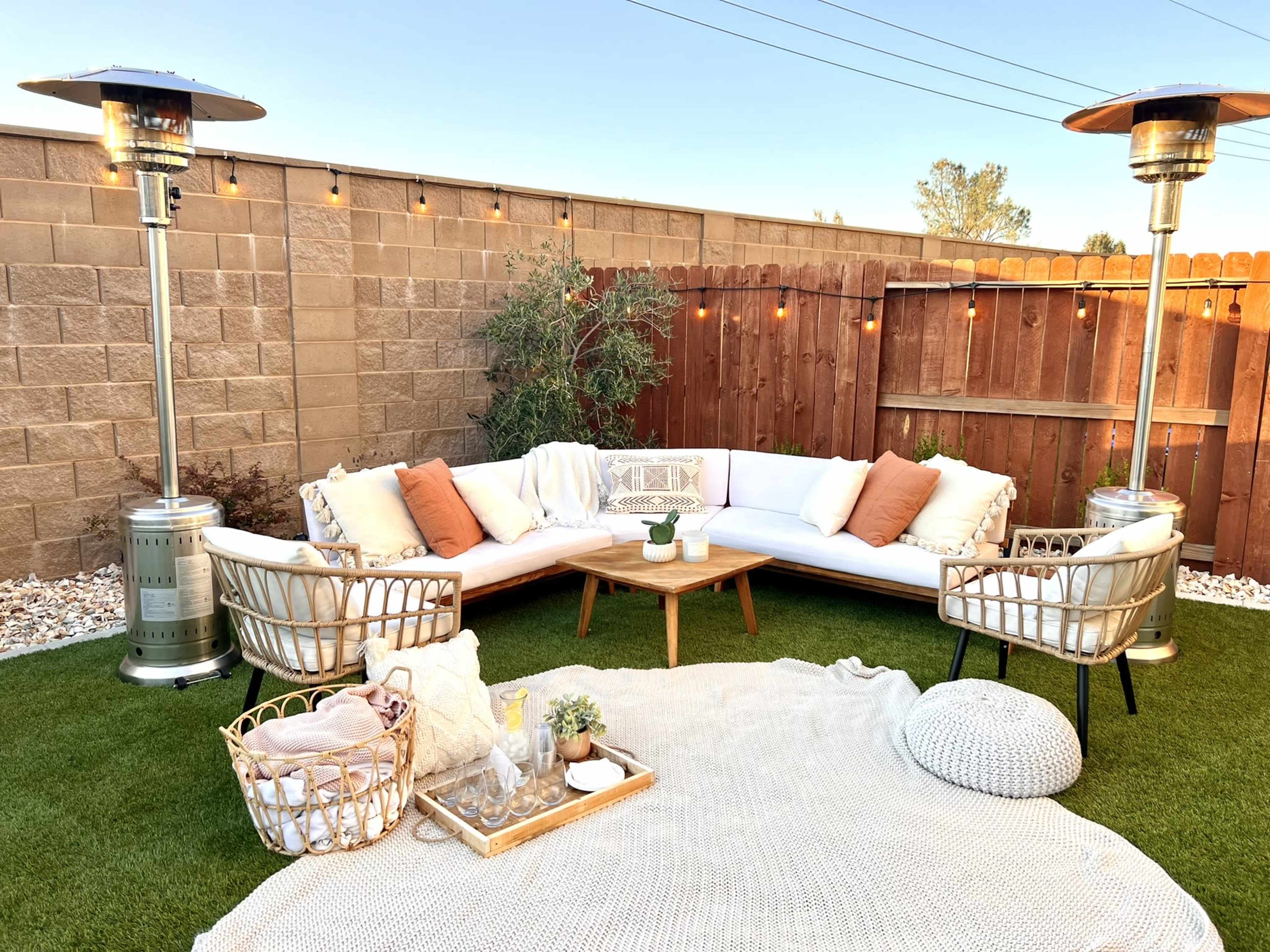 A cozy outdoor seating area features a sectional couch with throw pillows, a wooden coffee table, two chairs, and patio heaters, all surrounded by decorative lighting and greenery.