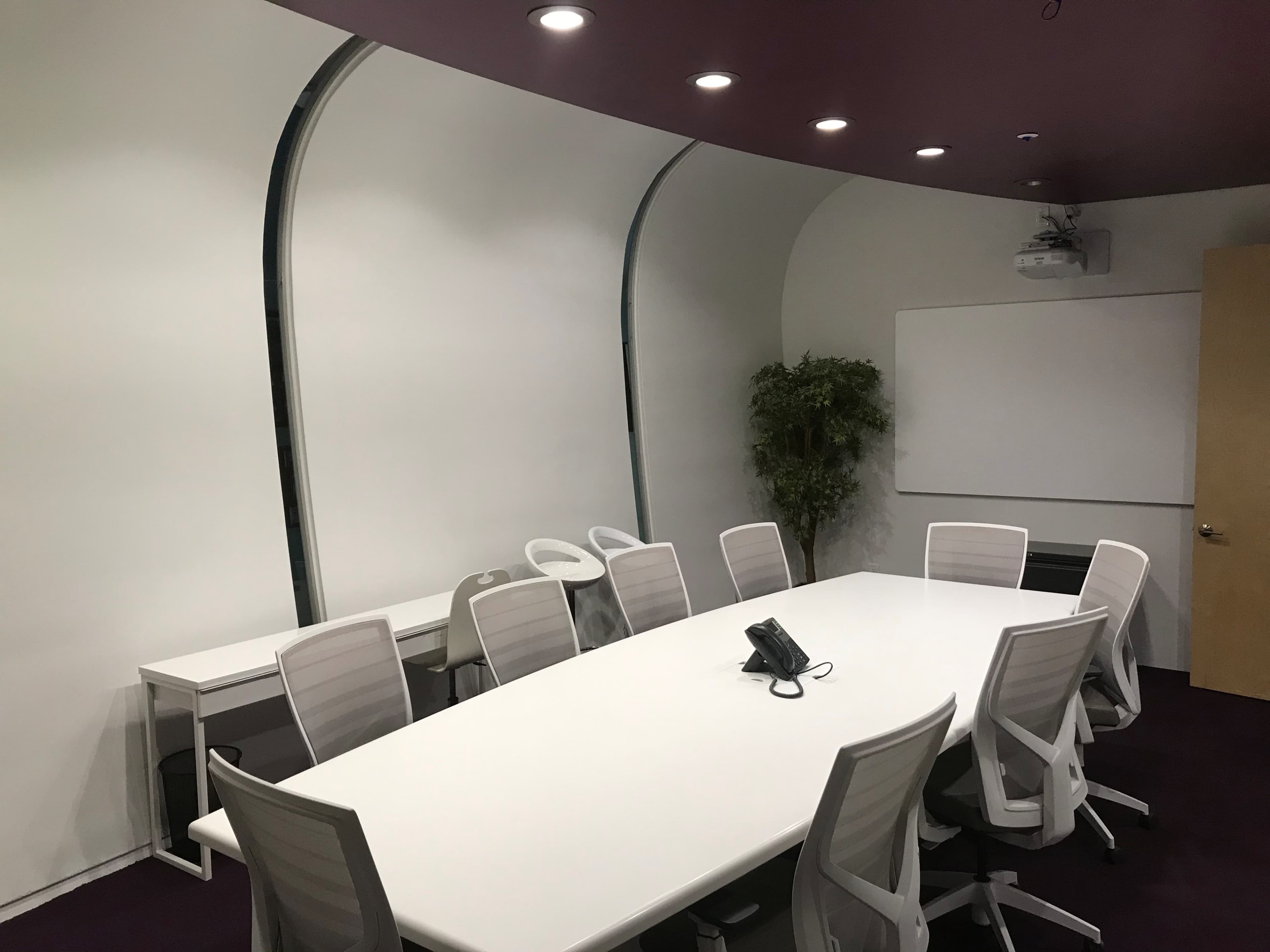 A modern conference room features a large white oval table surrounded by white chairs, with a projection screen and a potted plant in the corner.