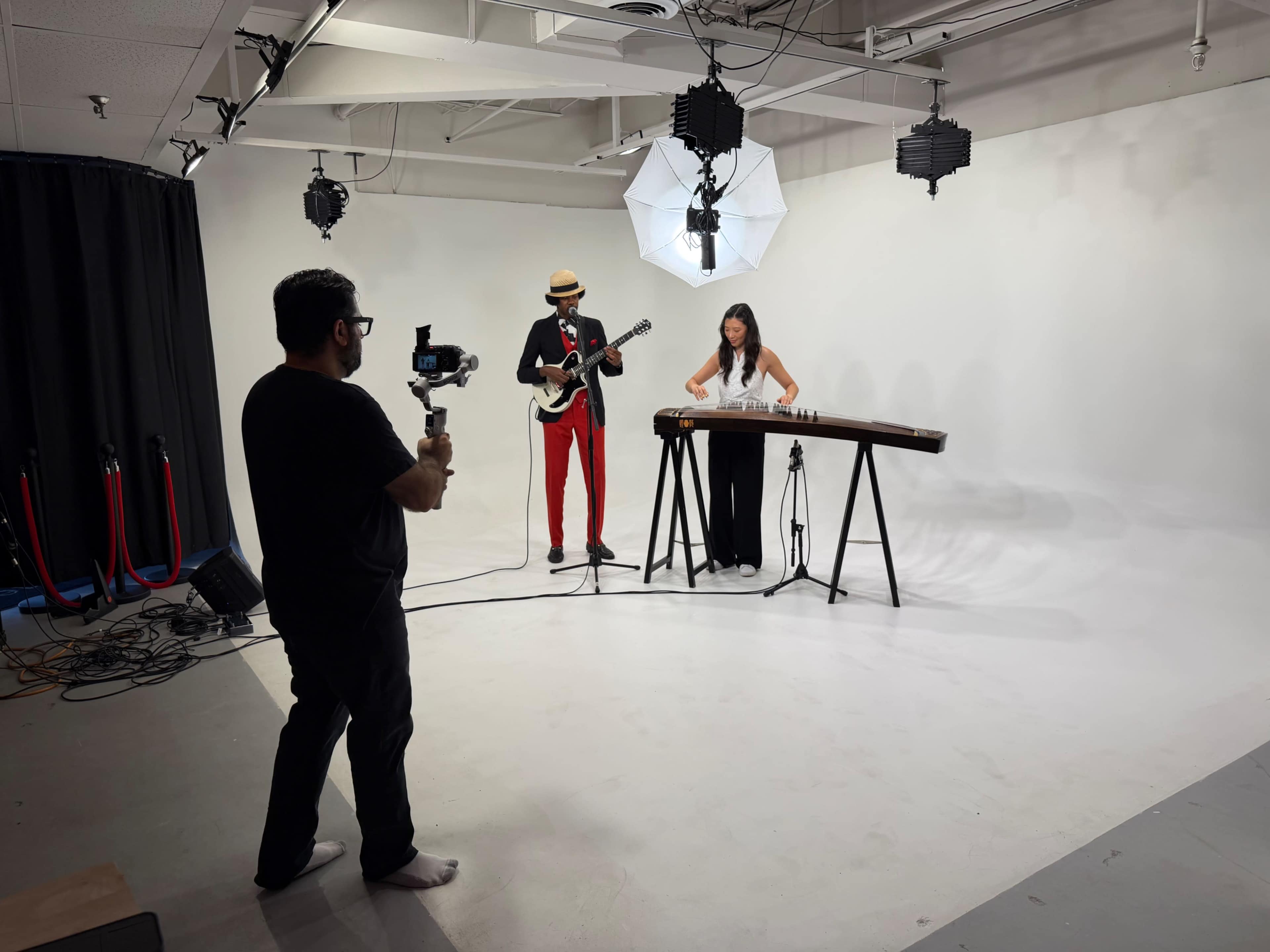 A musician in a red outfit plays a guitar while a woman in black performs on a stage instrument, as a camera operator records them in a white studio.
