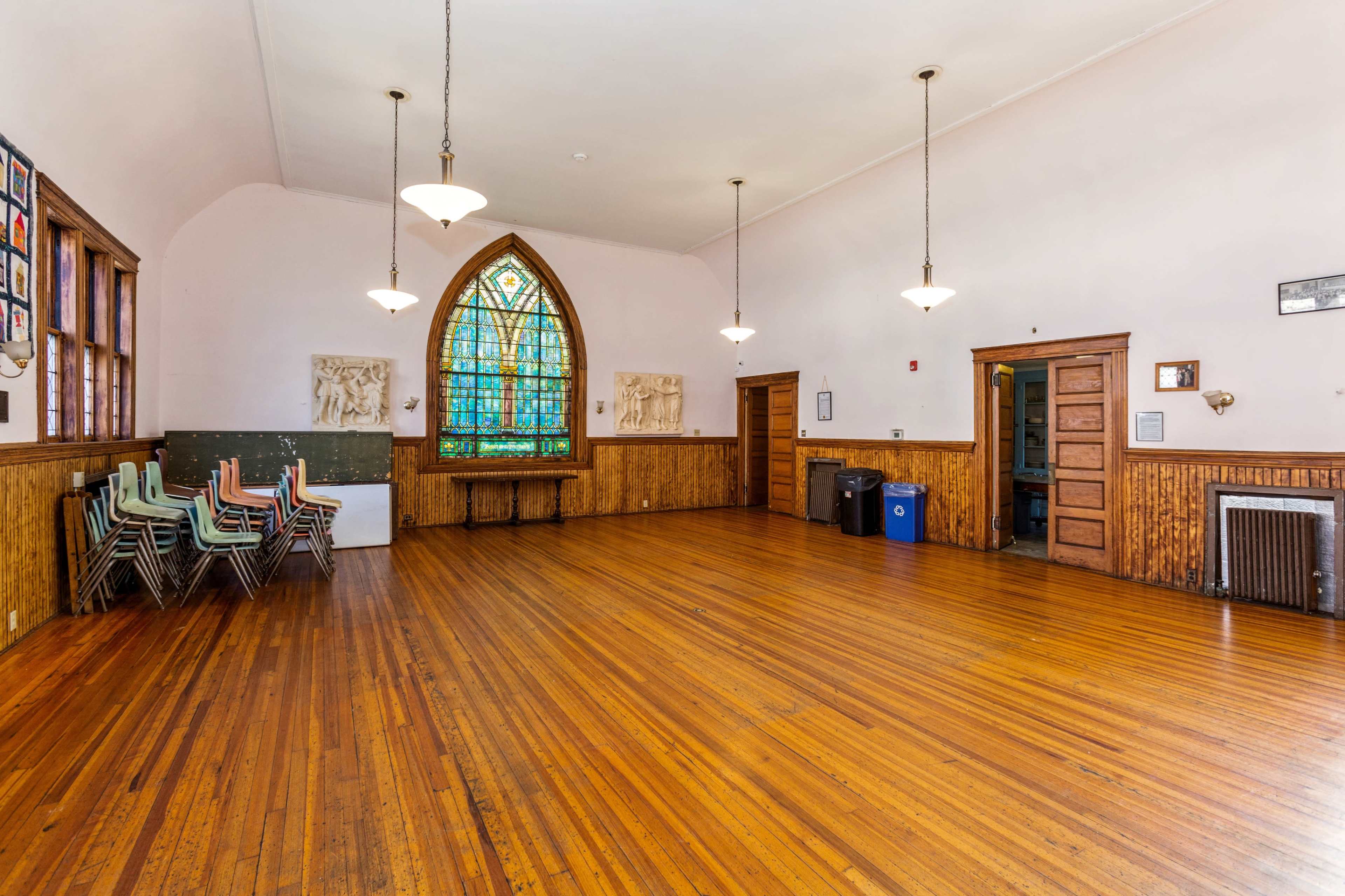 A spacious room with wooden floors, stained glass windows, and stacked chairs along one wall.