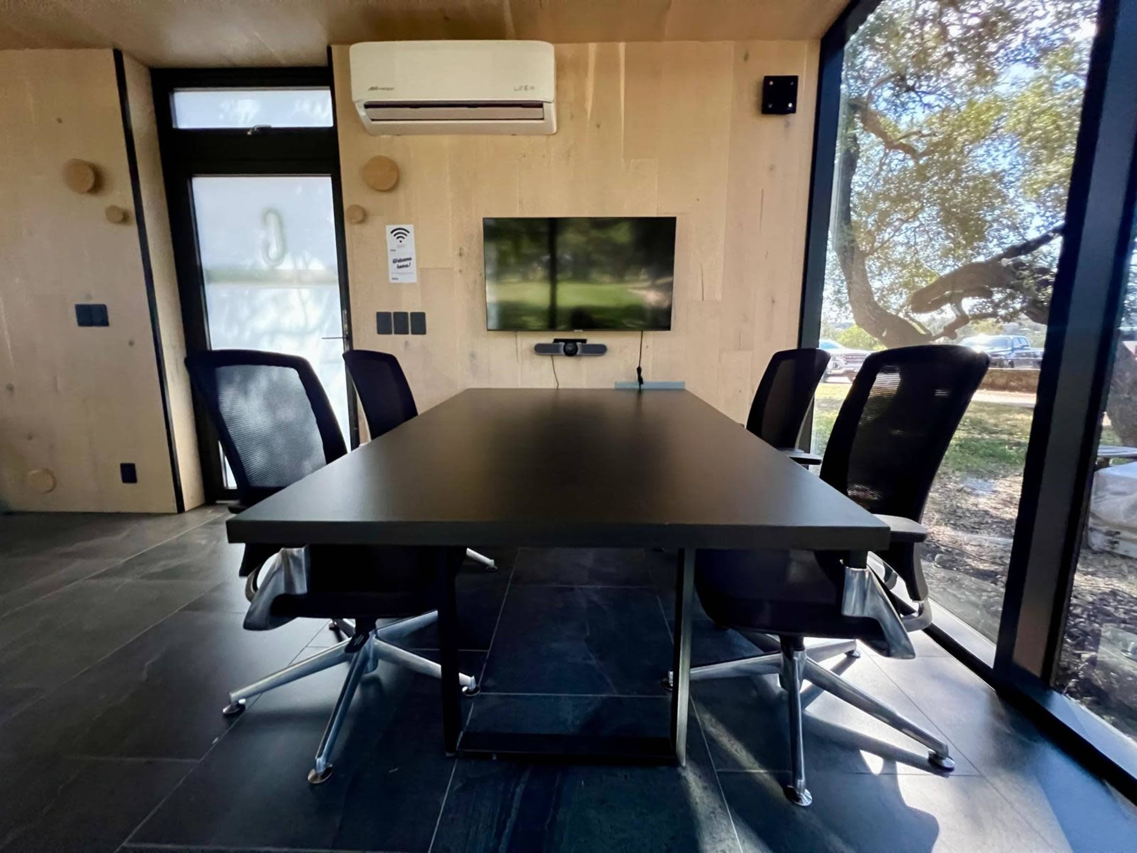A conference room features a wooden interior with a black table and four ergonomic chairs, accompanied by a wall-mounted TV and a large window overlooking trees.