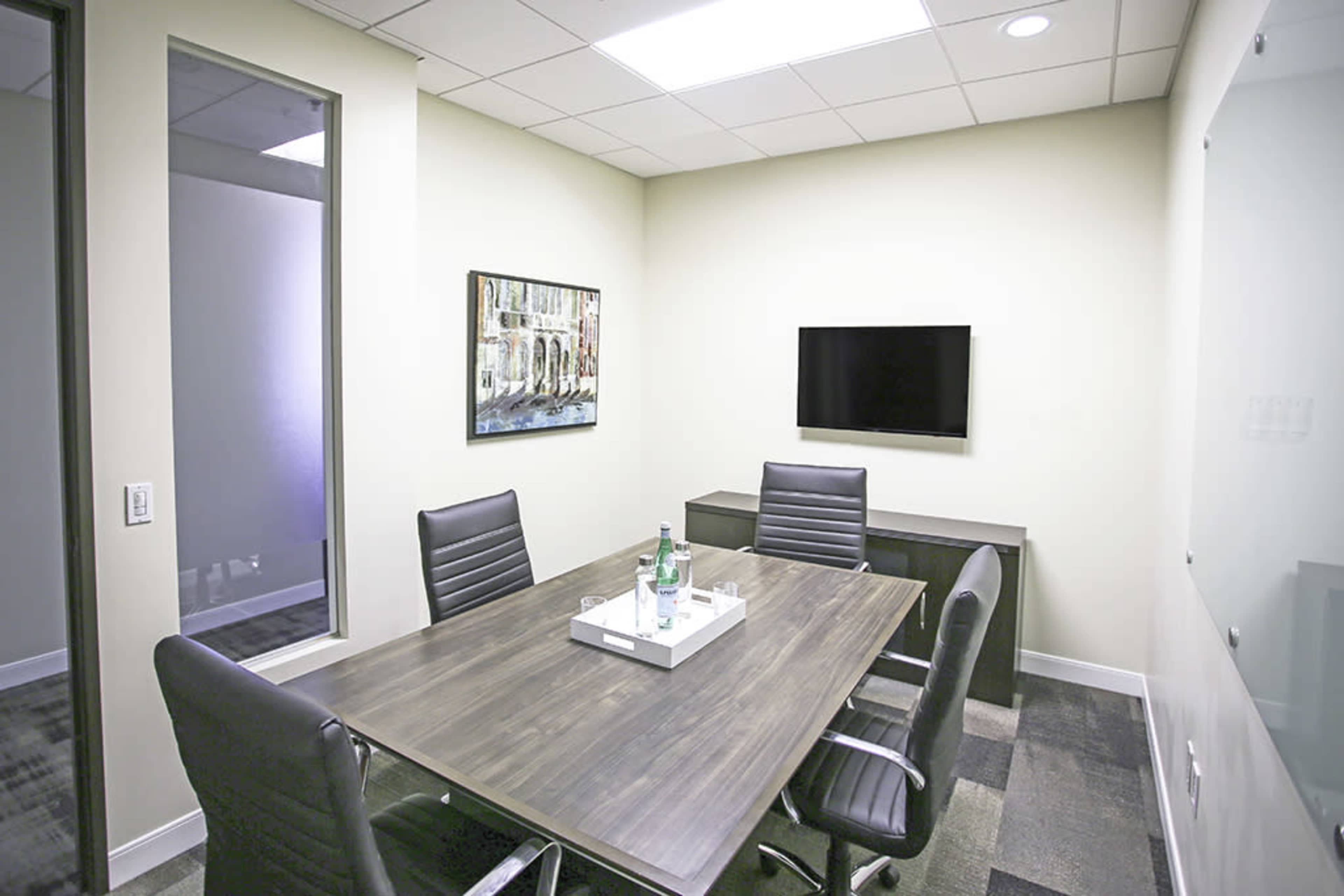 A small meeting room with a rectangular table, black leather chairs, a television mounted on the wall, and a minimalist decor.