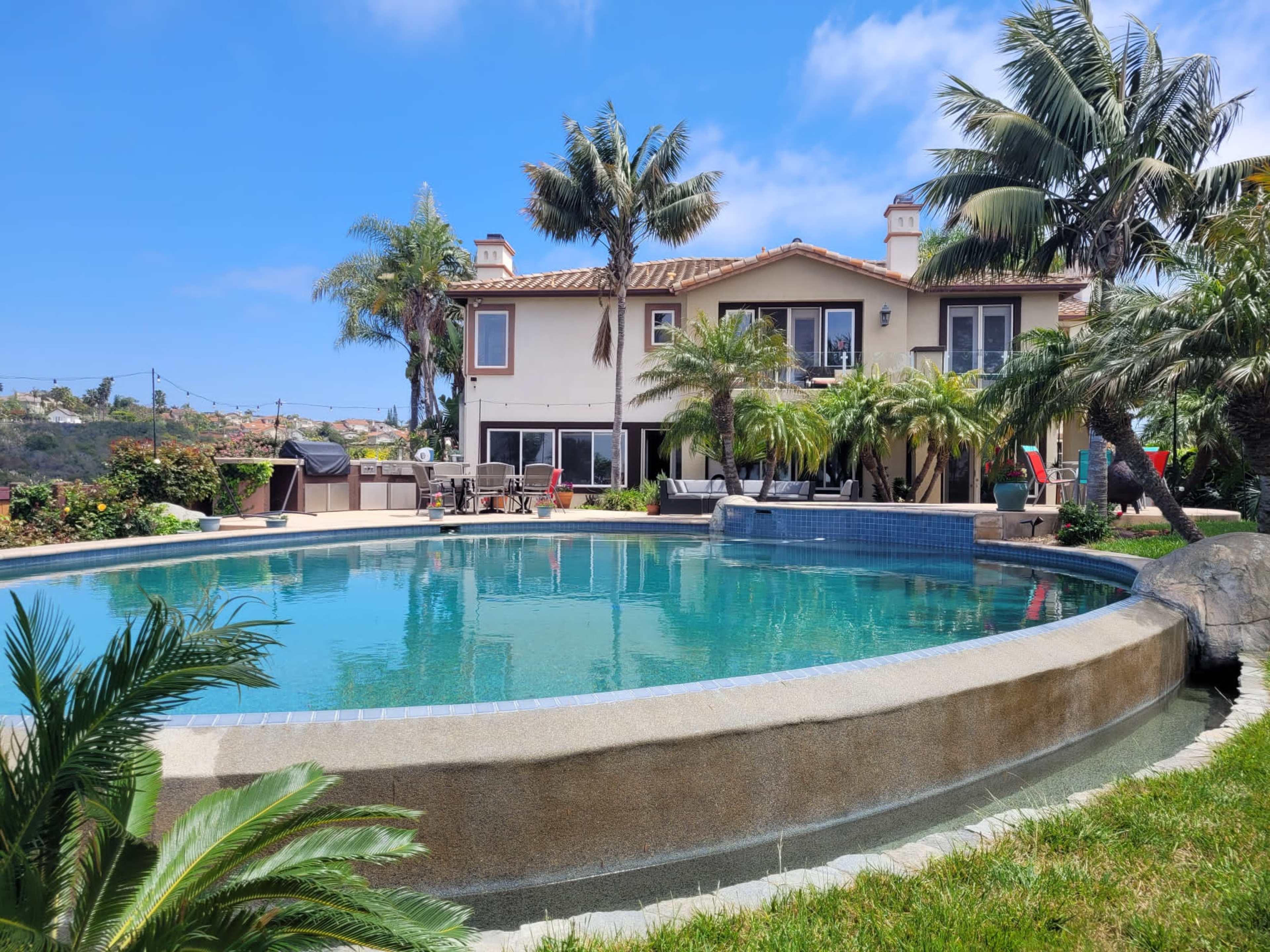 A large swimming pool surrounded by palm trees and a landscaped garden adjoins a multi-level house with a balcony in the background.