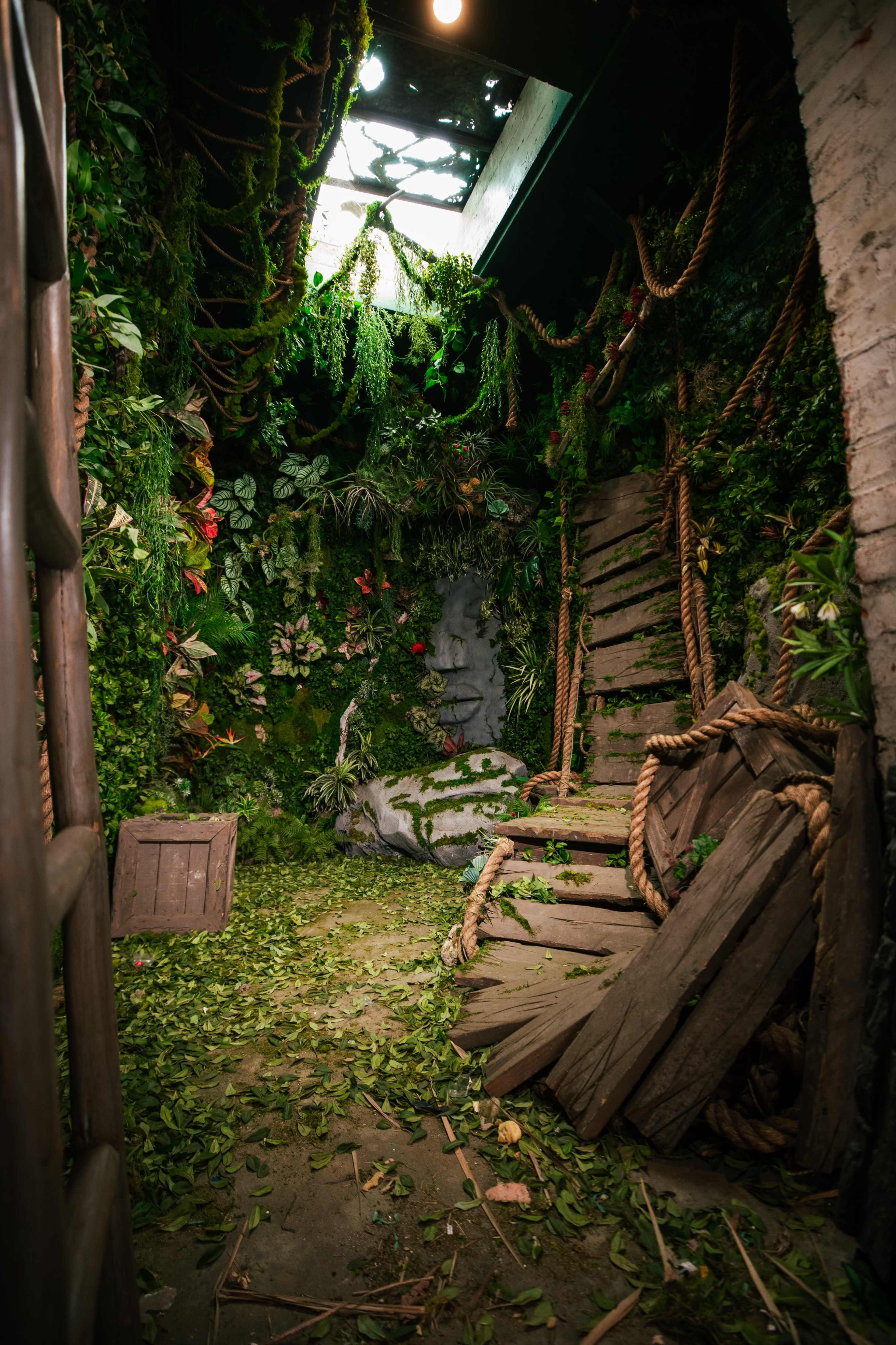 An indoor space overgrown with plants and vines, featuring a partially broken wooden swing and a crate amidst fallen leaves.