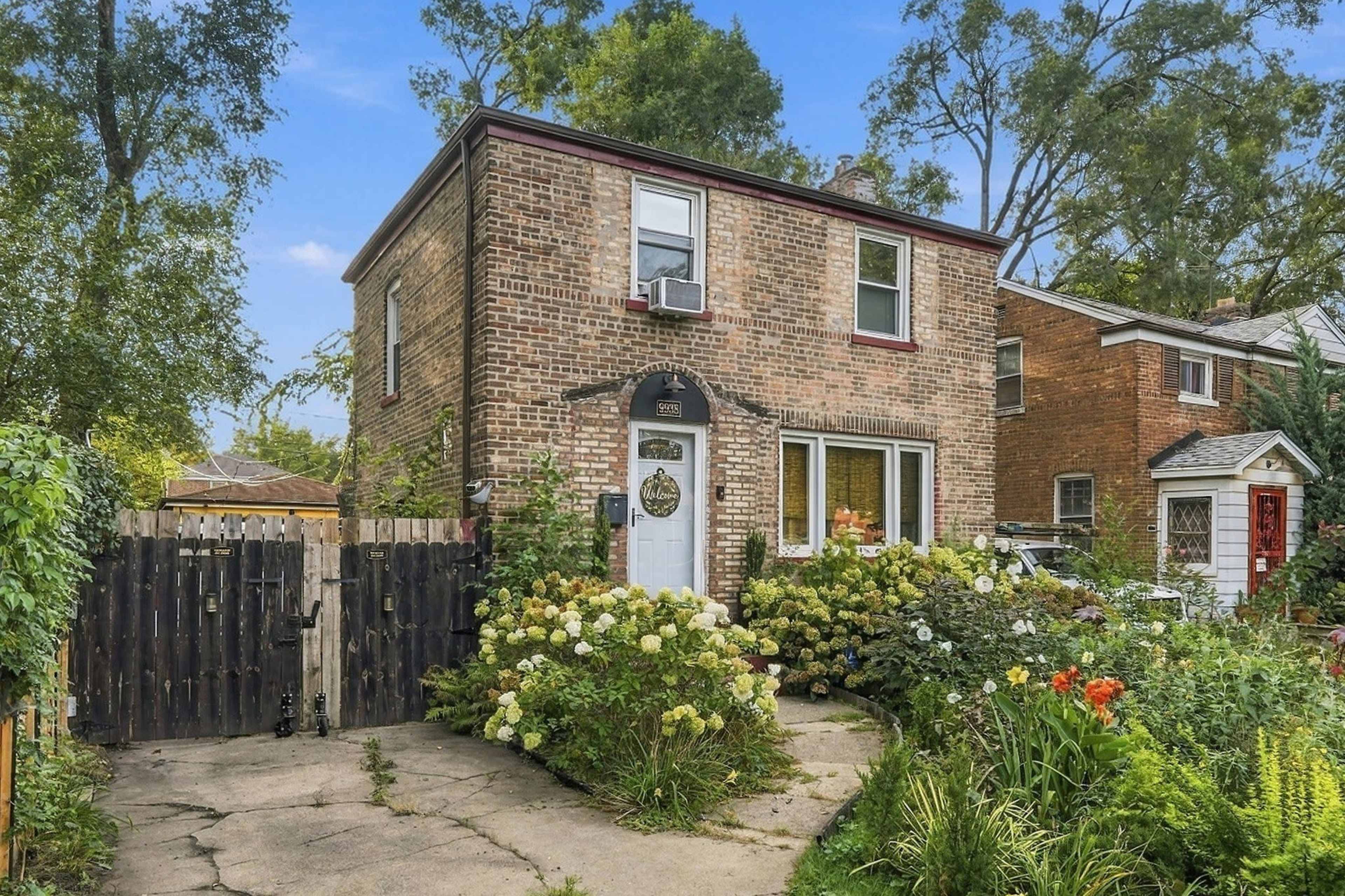 A two-story brick house with a front garden featuring various blooming plants and a gravel driveway is pictured.