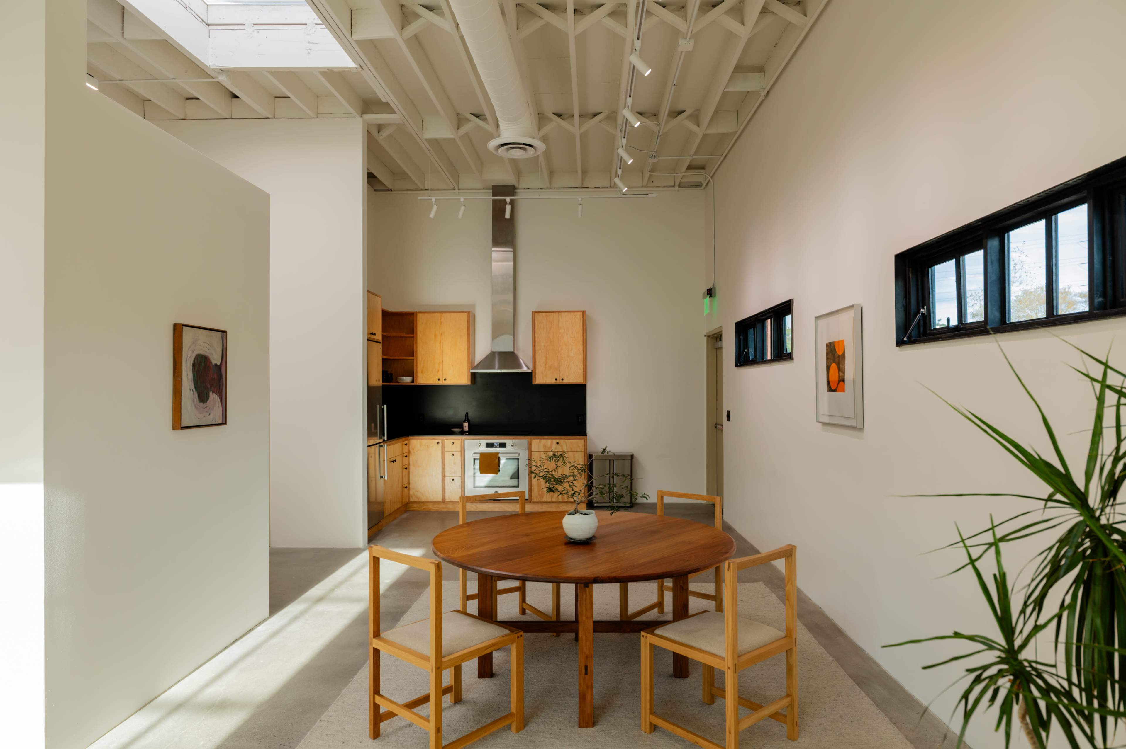A minimalist kitchen and dining area features a round wooden table surrounded by four chairs, with a simple kitchen setup in the background and a potted plant to the right.