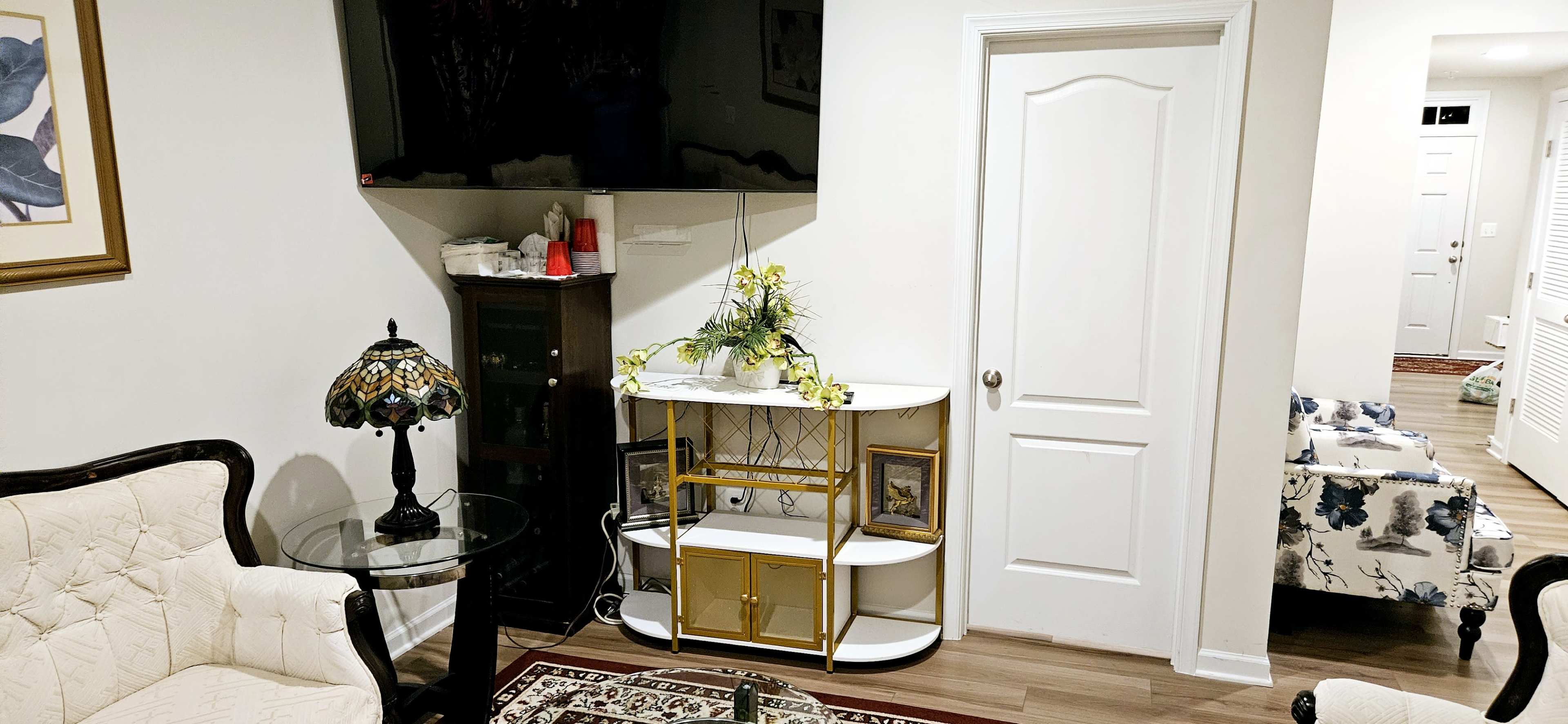 The image shows a living room with a television mounted on the wall, a decorative table with a lamp, and a white cabinet against the wall, with a door leading to another room in the background.